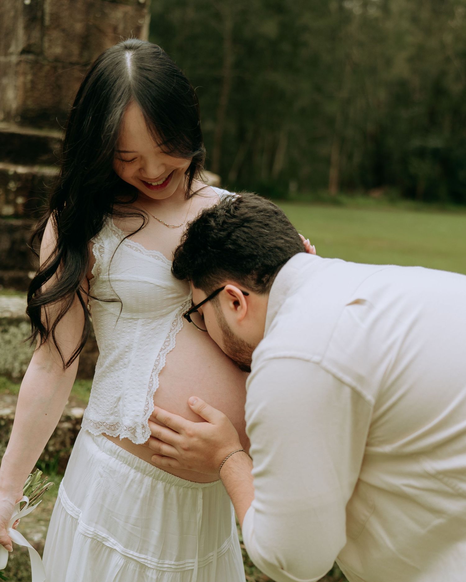 Partner kissing pregnant woman's bare belly during an outdoor maternity photo session in a park.