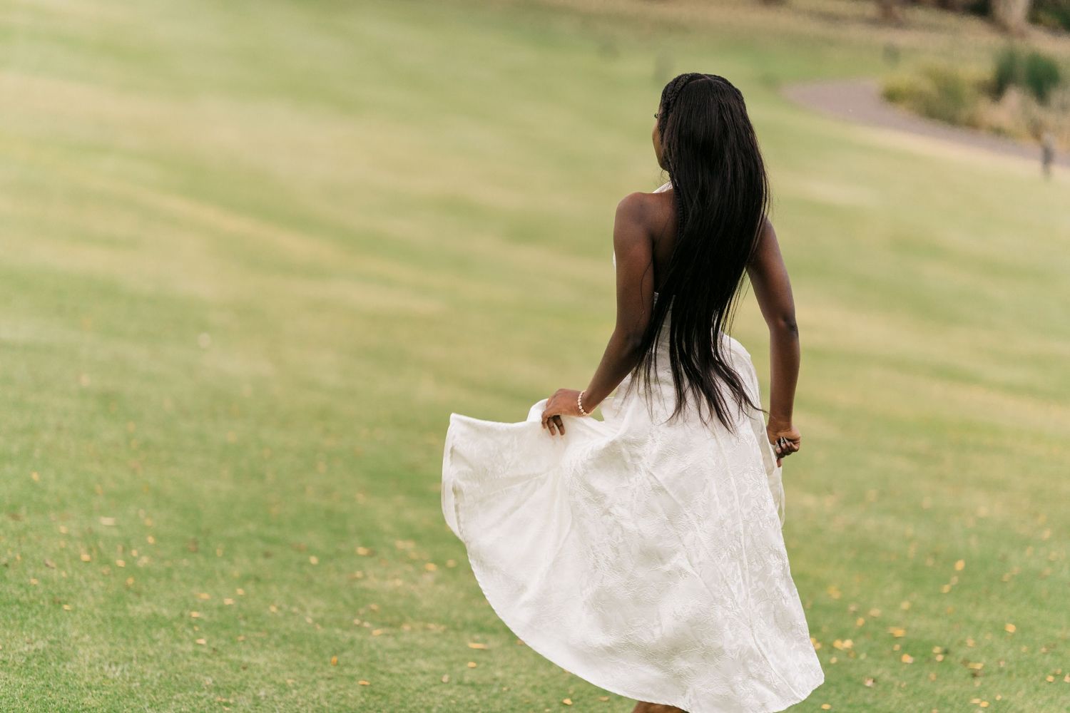 A young Black woman in a flowing white midi dress walks across a green lawn, viewed from behind.