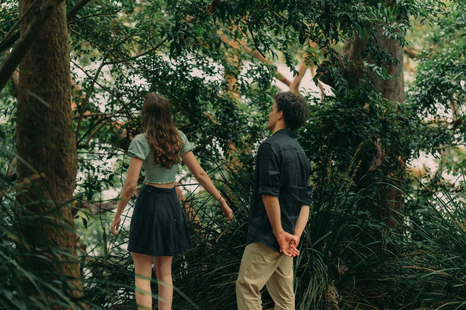 A couple holding hands while walking through a dense green forest from behind.