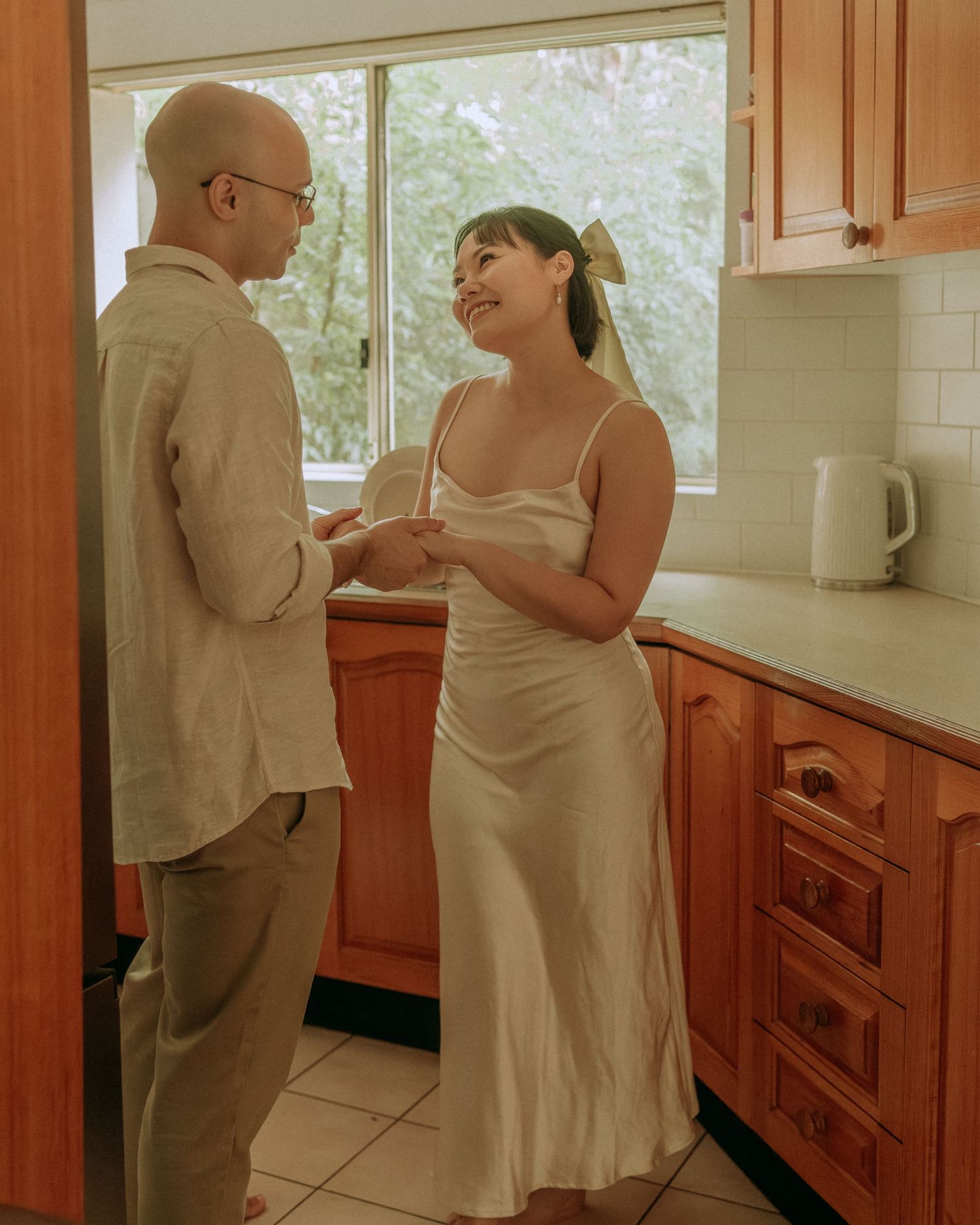 Couple smiling and holding hands in a bright kitchen, woman in elegant satin slip dress with bow.