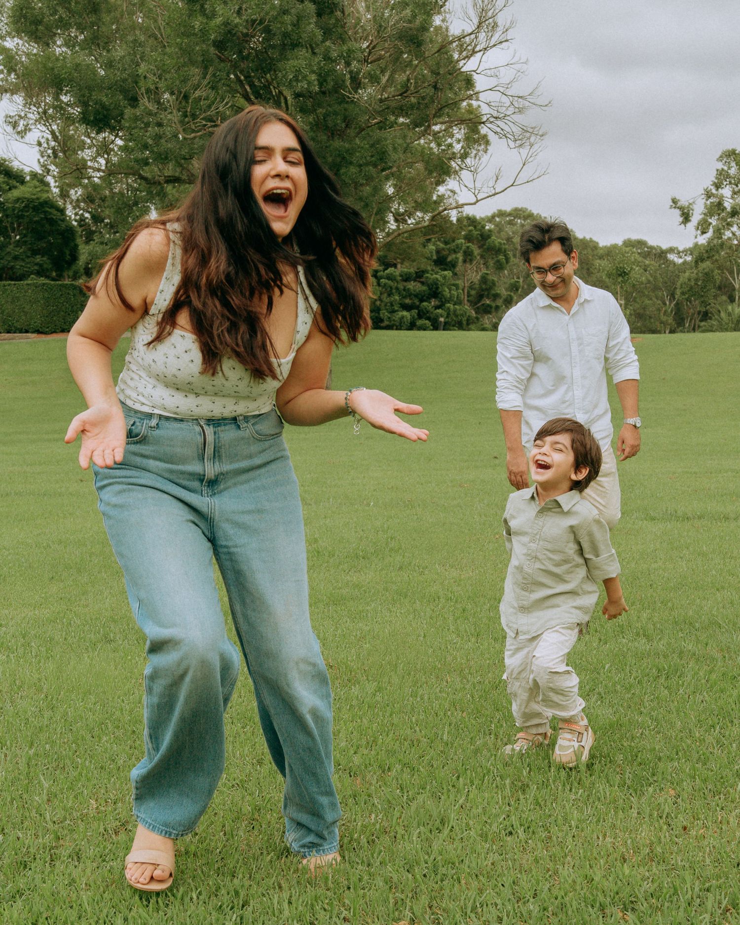 Laughing mother and toddler playing on green lawn while father watches warmly in background.