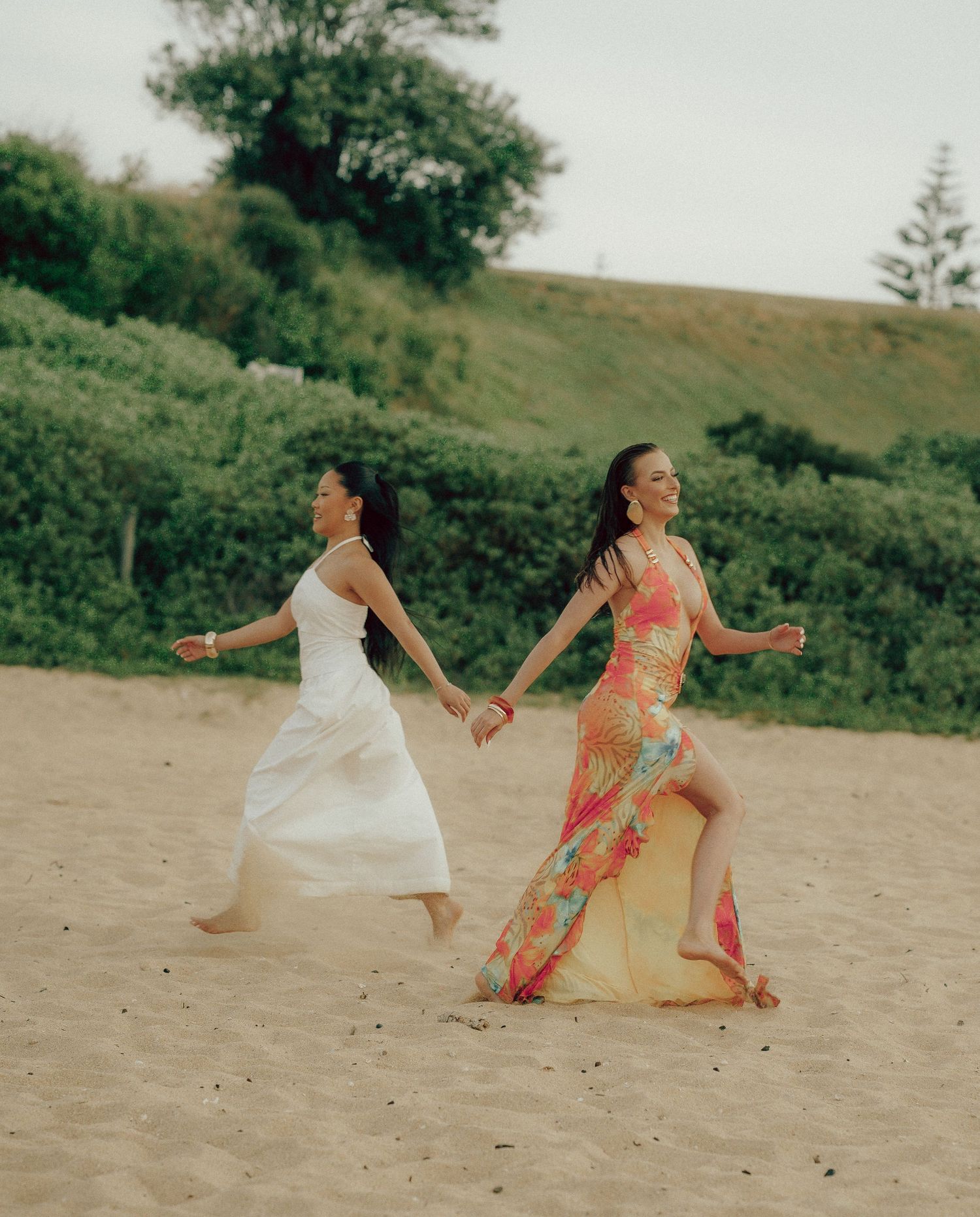 Two women holding hands running on sandy beach, one in white dress, one in colorful floral gown.