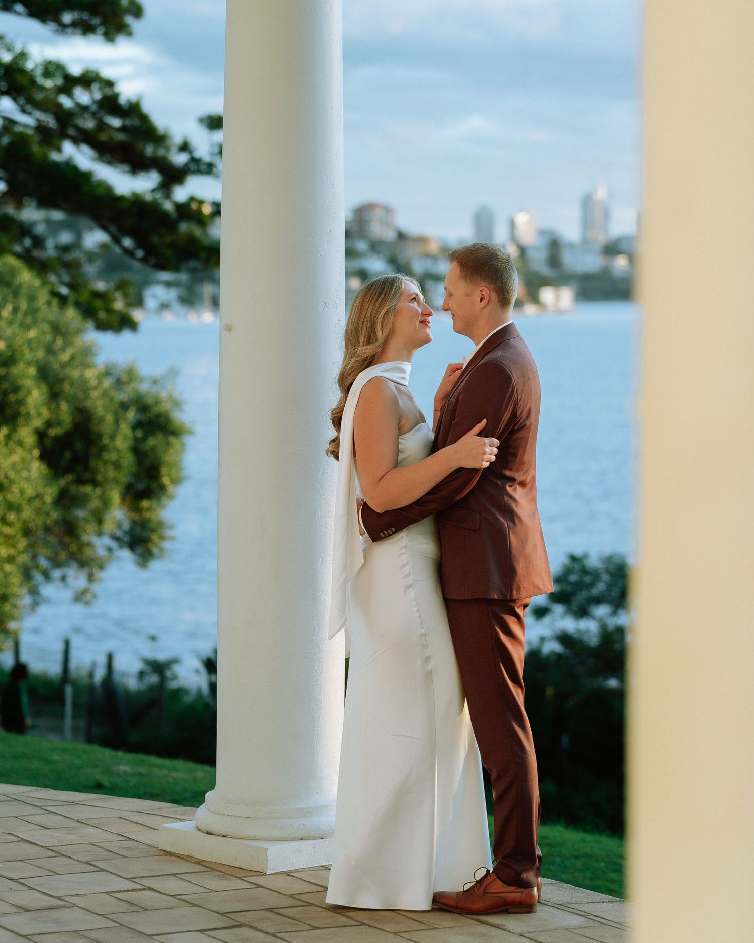 Newlyweds embrace between white columns with Sydney harbour cityscape and water in background.