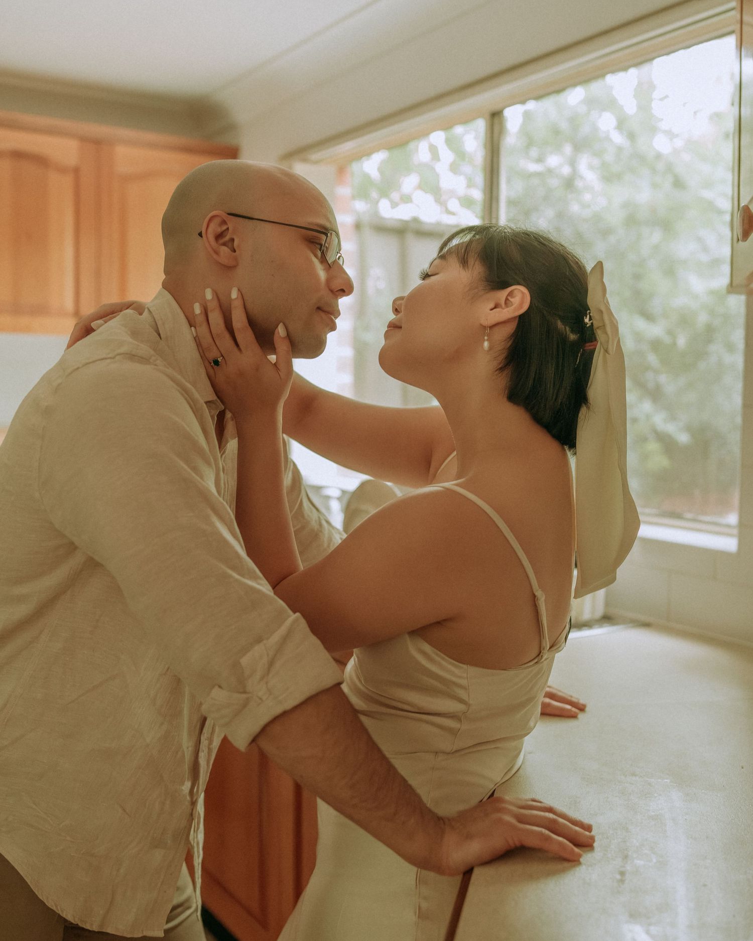 Couple in romantic moment, woman cupping mans face leaning in for a kiss in sunlit kitchen.