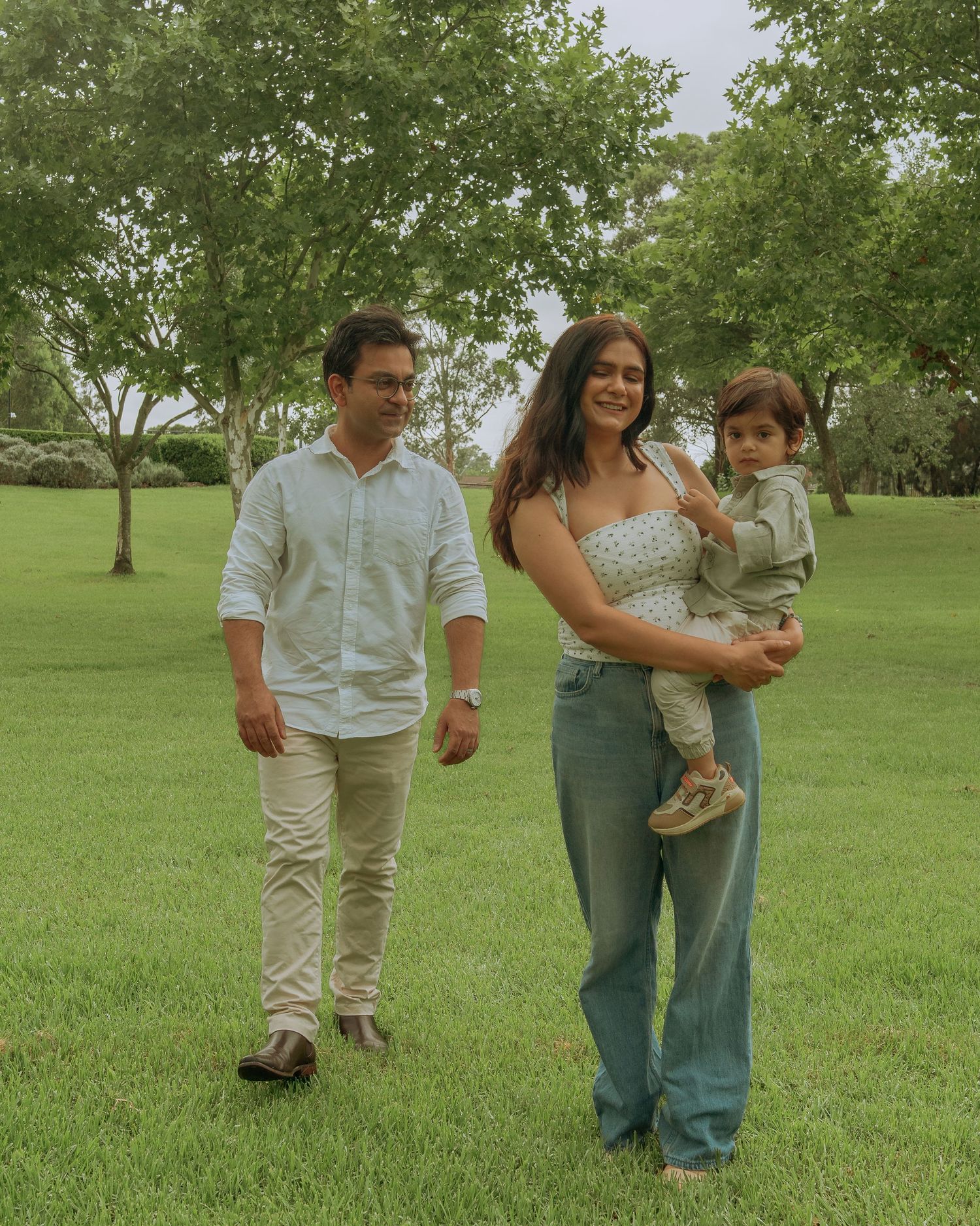 Family of three walking on green grass, mother holding toddler while father strolls beside them in a park.
