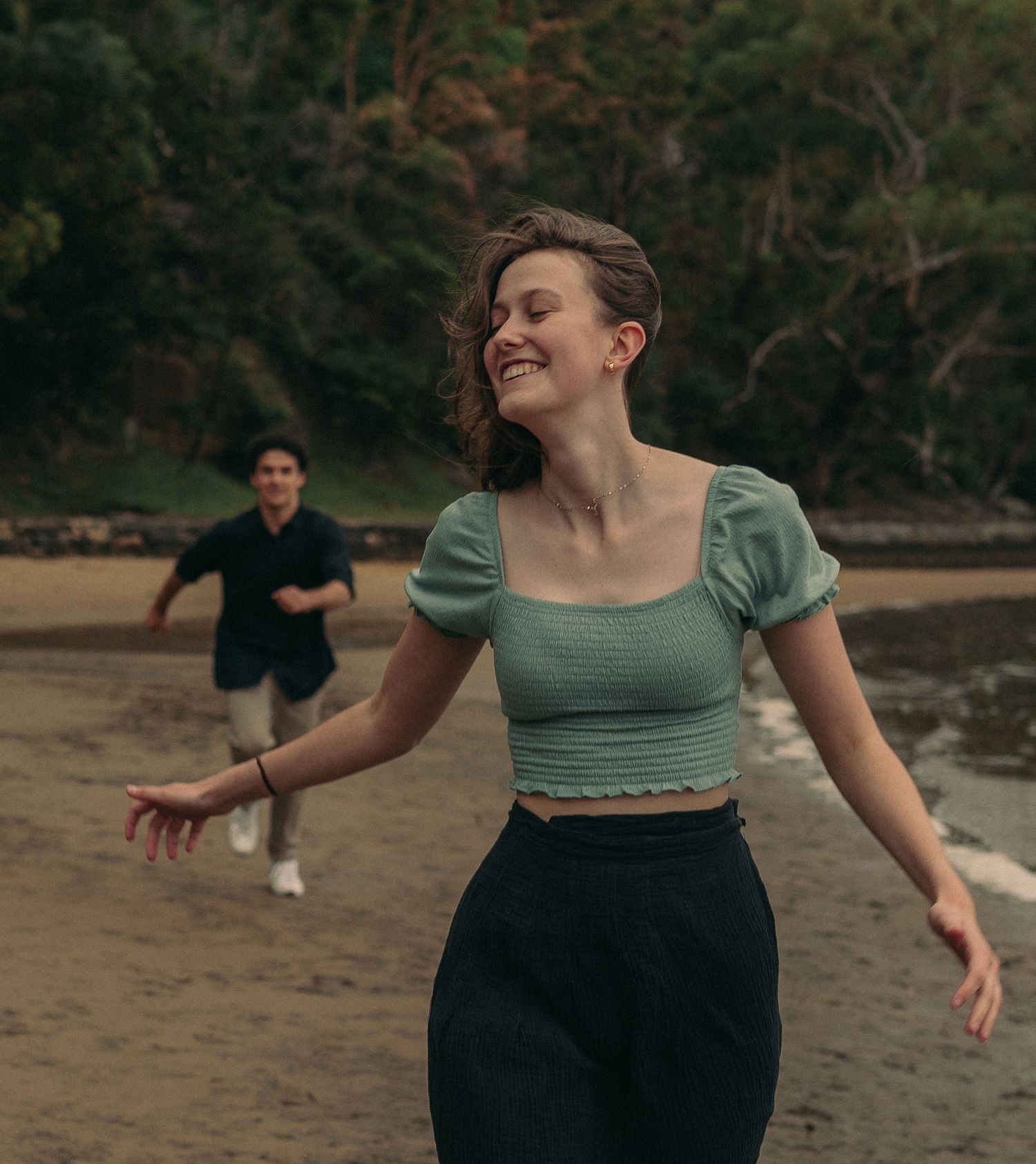Joyful woman in a green crop top running on a beach with a man following behind.