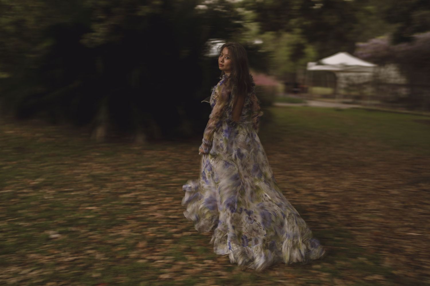Woman in flowing floral gown spinning in a lush green garden at dusk.