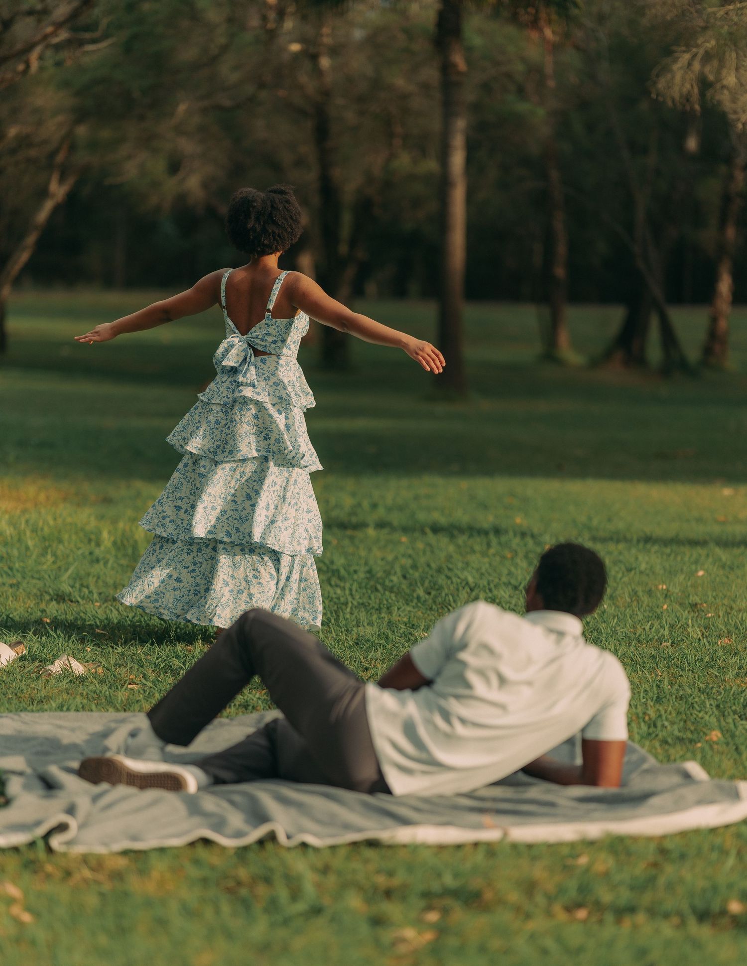 A woman in a tiered floral dress spins with arms outstretched near a man lying on a park blanket.