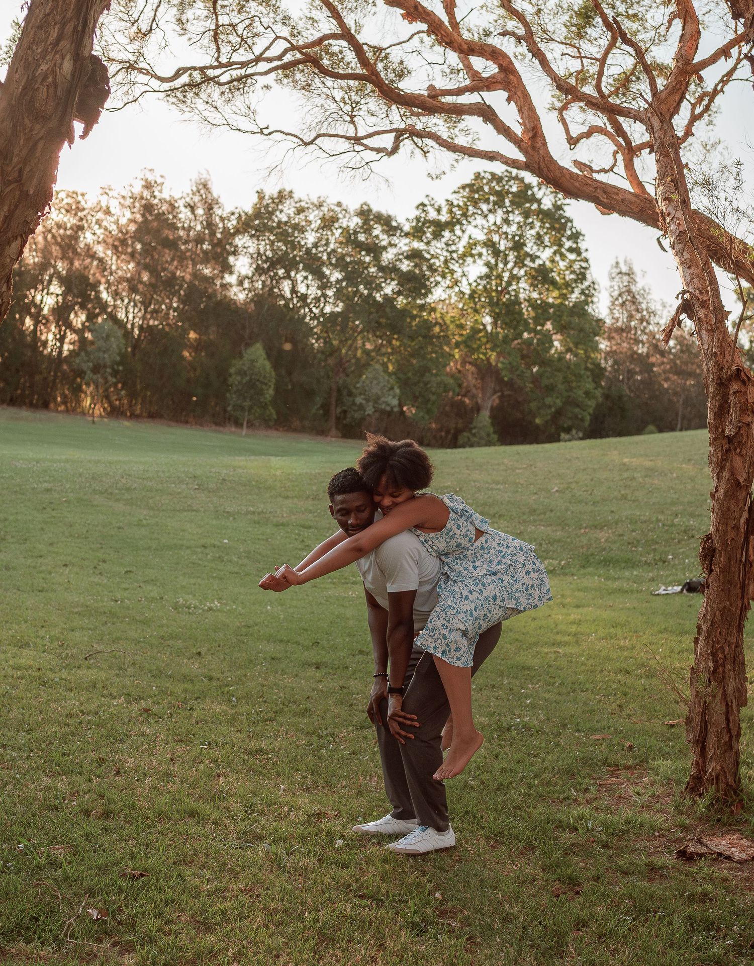 A woman joyfully jumps on her partners back near a large tree in a sunny green park.