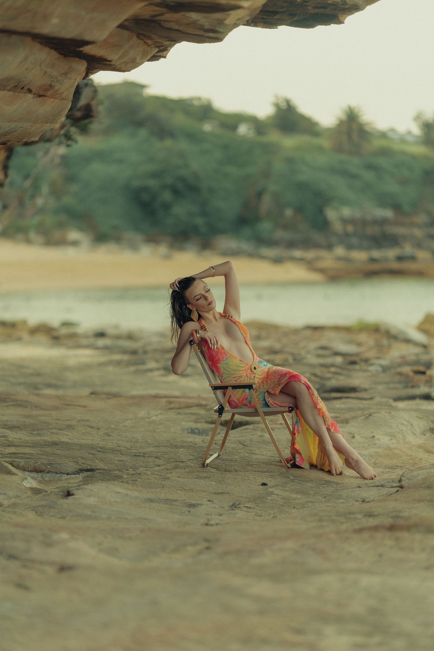 Woman in vibrant floral dress relaxing on folding chair on rocky beach with coastal scenery behind.