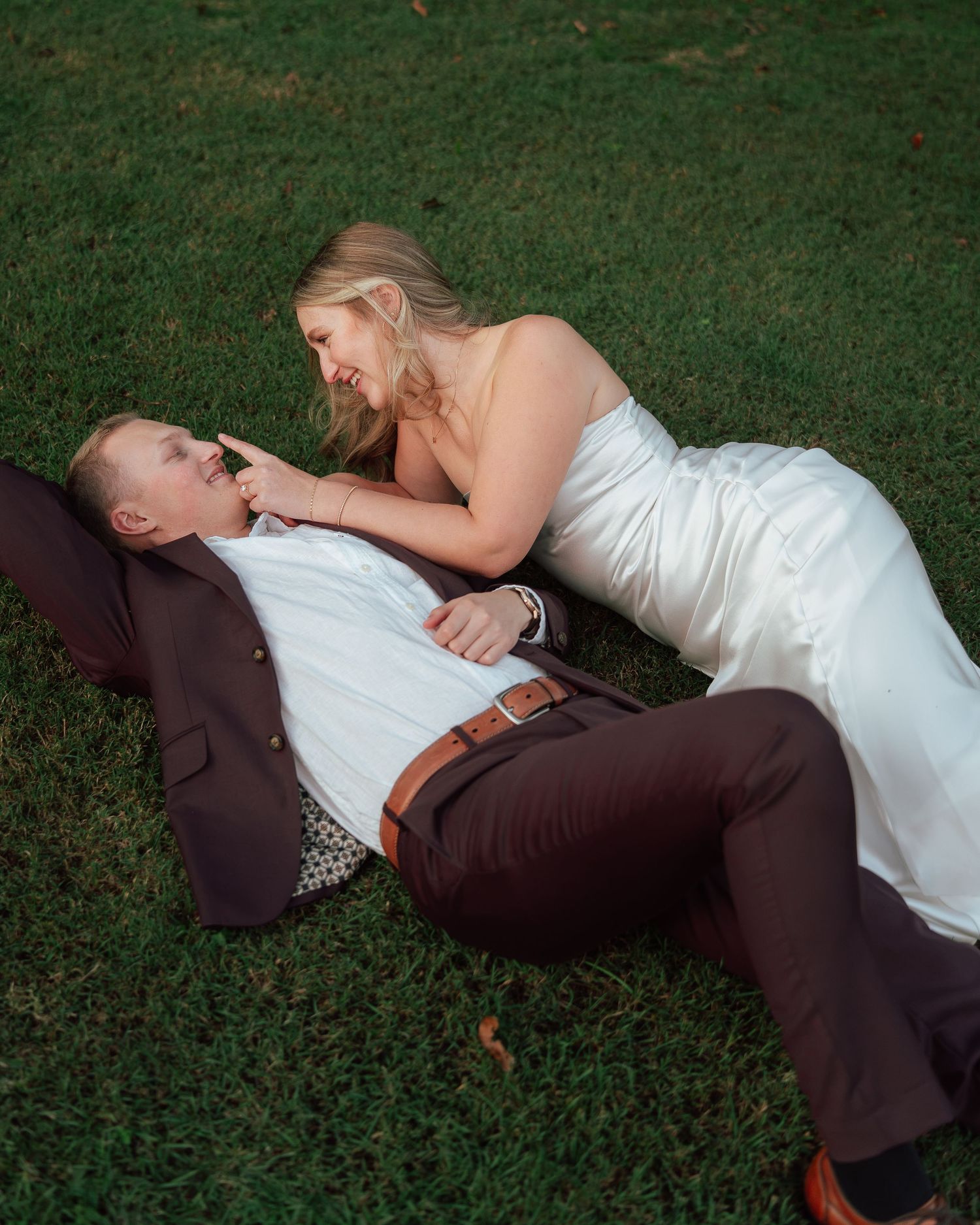 Laughing bride in white gown leans over groom lying on lush green grass during wedding shoot.