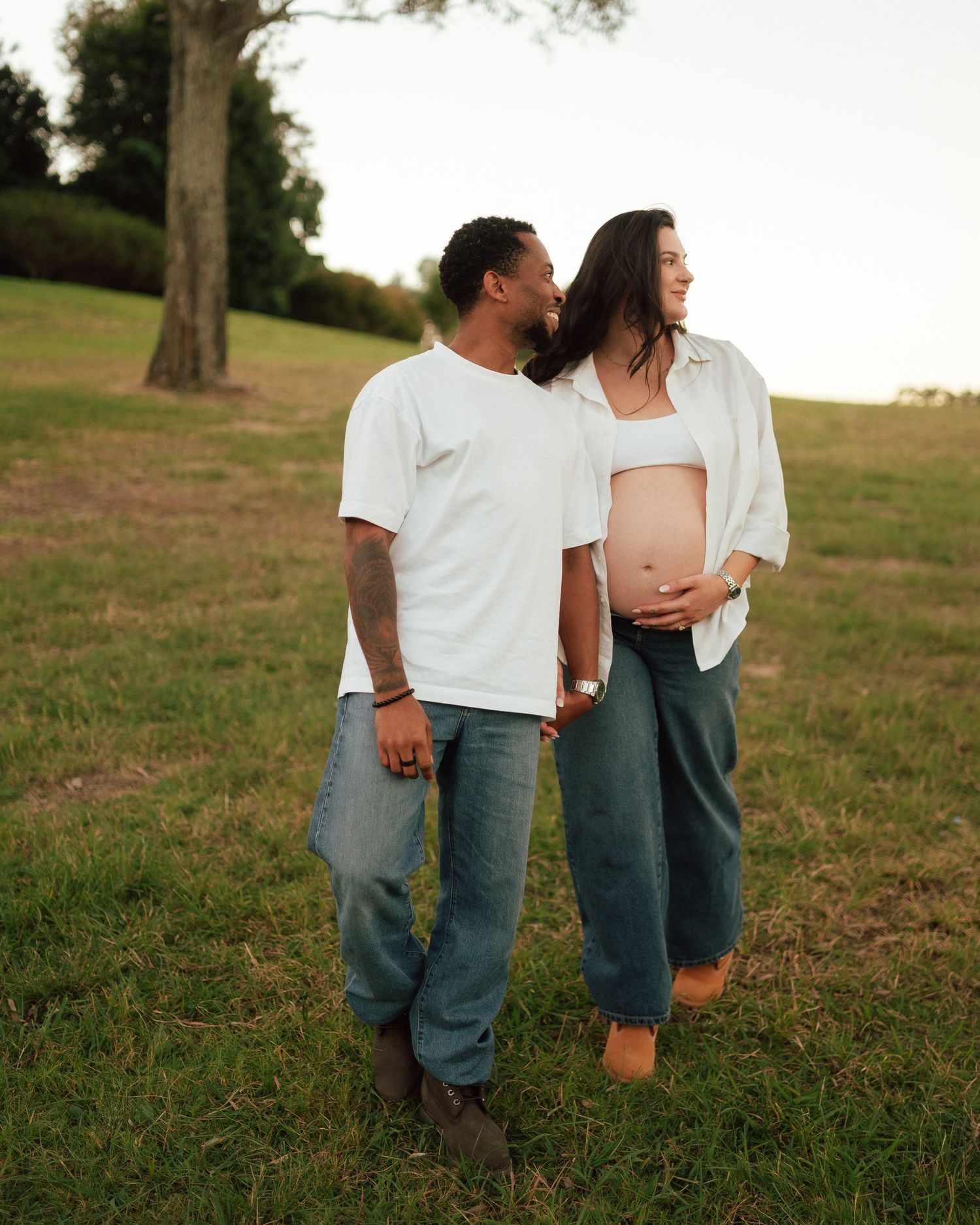 Couple standing in a green field at golden hour, man embracing pregnant woman from behind lovingly.