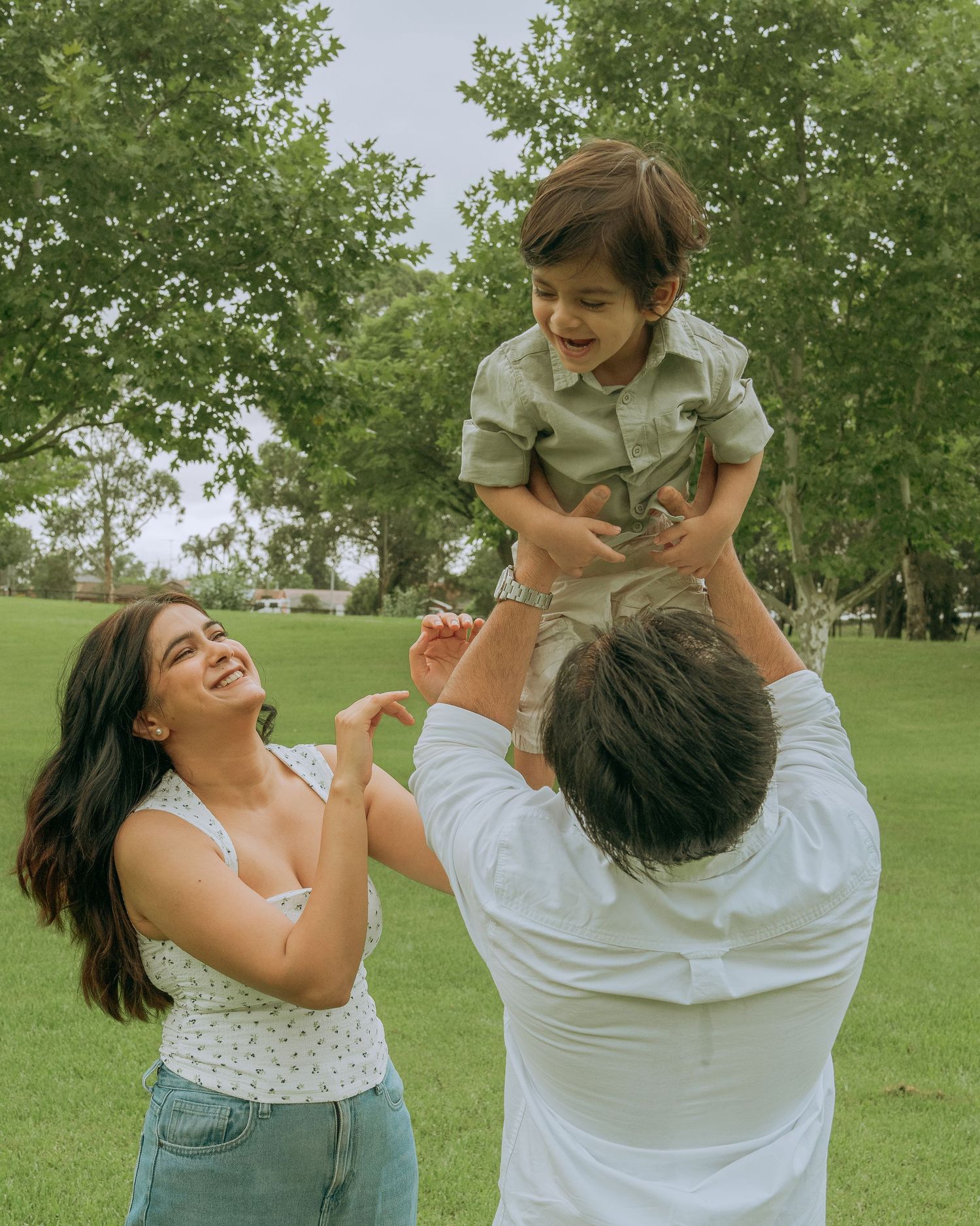 Joyful parents lifting laughing toddler in the air on a green lawn.
