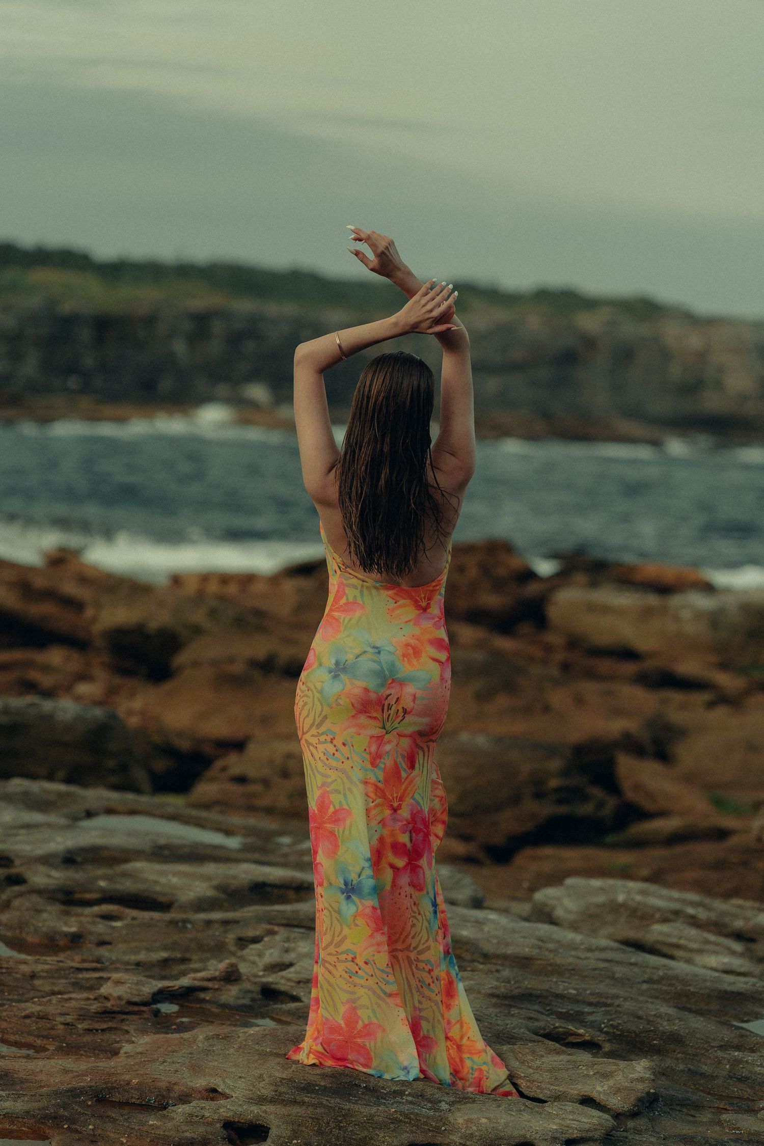 Woman in vibrant floral maxi dress standing on coastal rocks with arms raised.