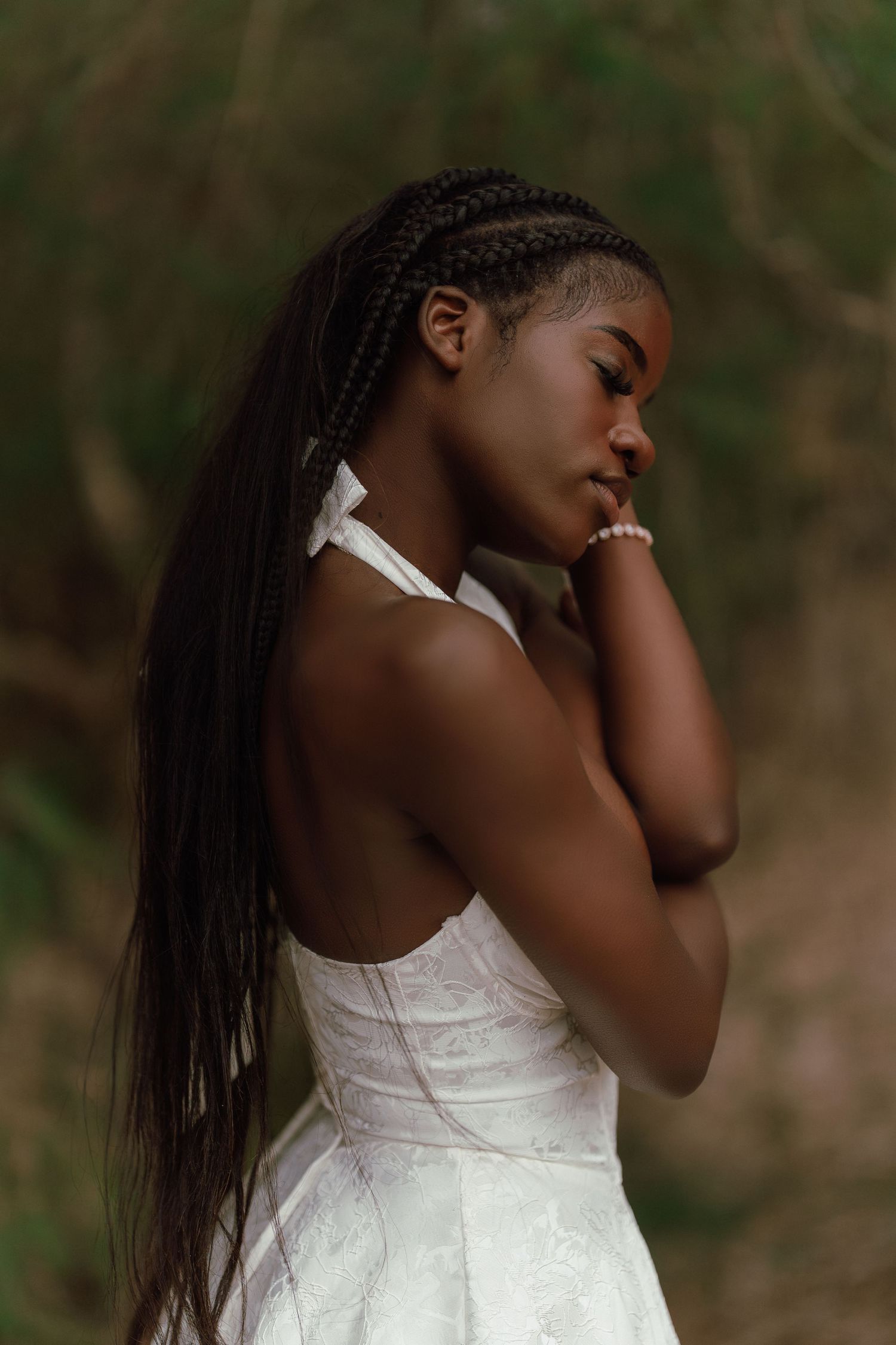 Young Black woman in white halter dress with long braids poses with eyes closed in natural setting.