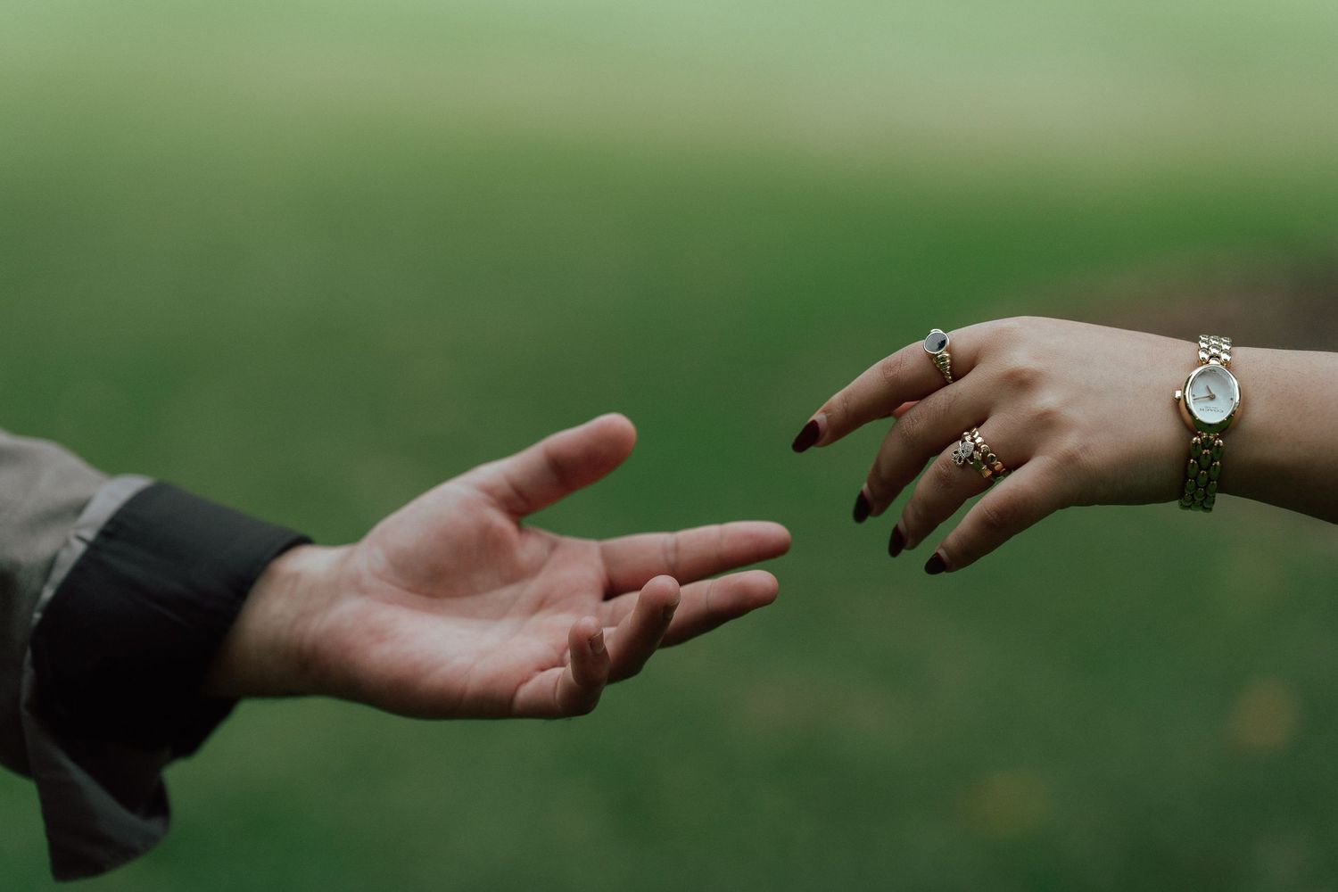 Close-up of two hands reaching toward each other with gold rings and watch on green background.