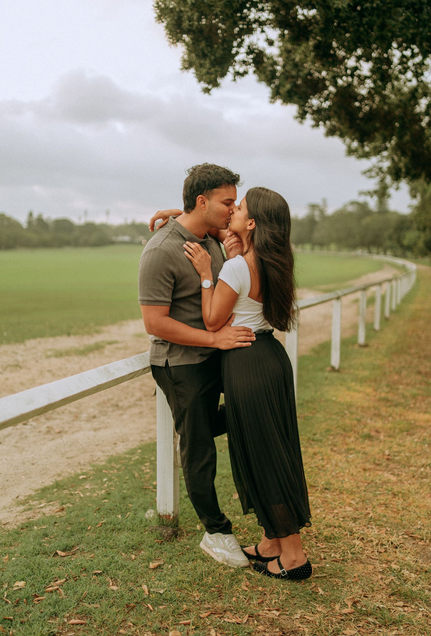 A couple shares a romantic kiss while leaning against a white fence along a green park pathway.