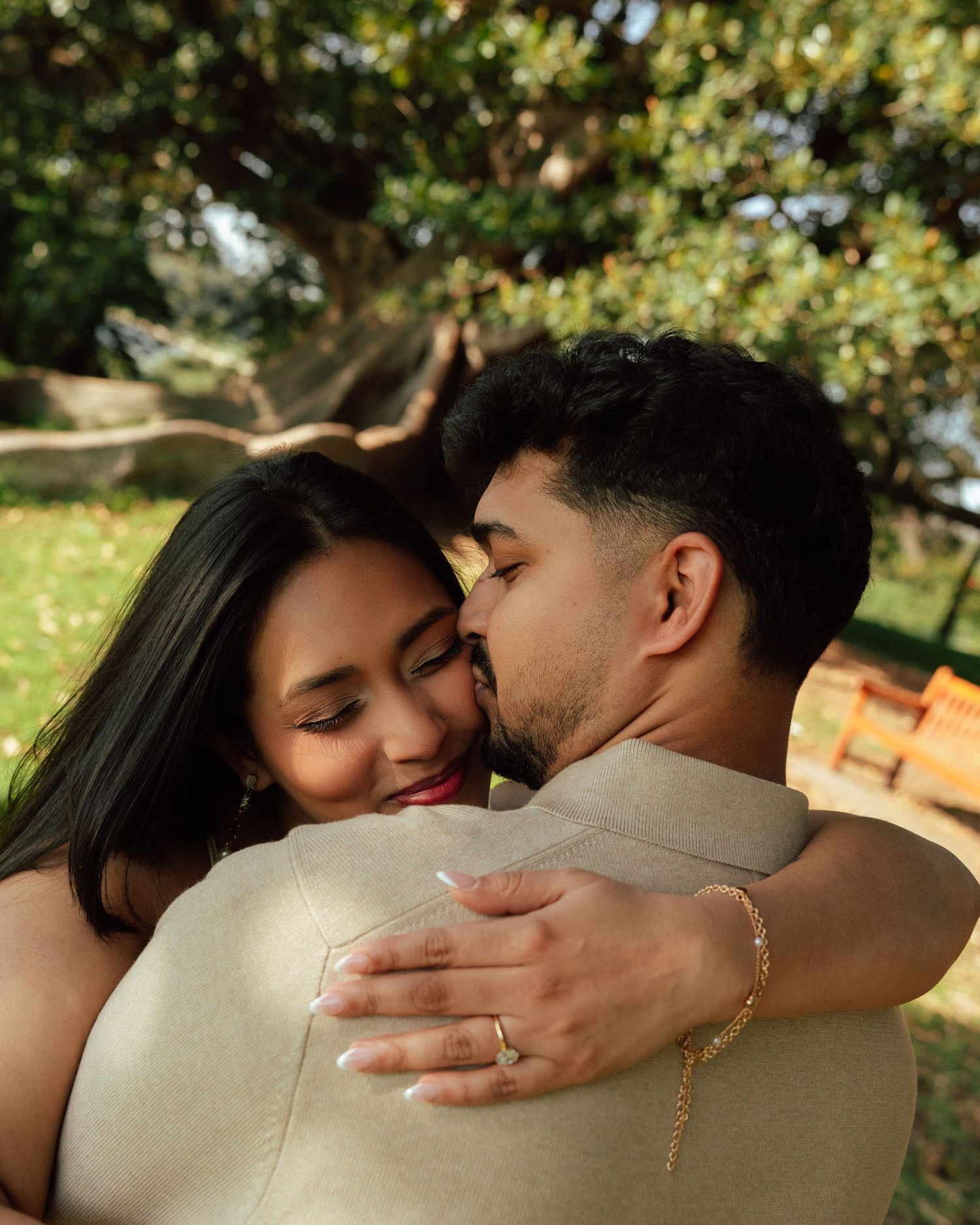 A couple shares a tender embrace outdoors, woman smiling with an engagement ring visible, trees in background.