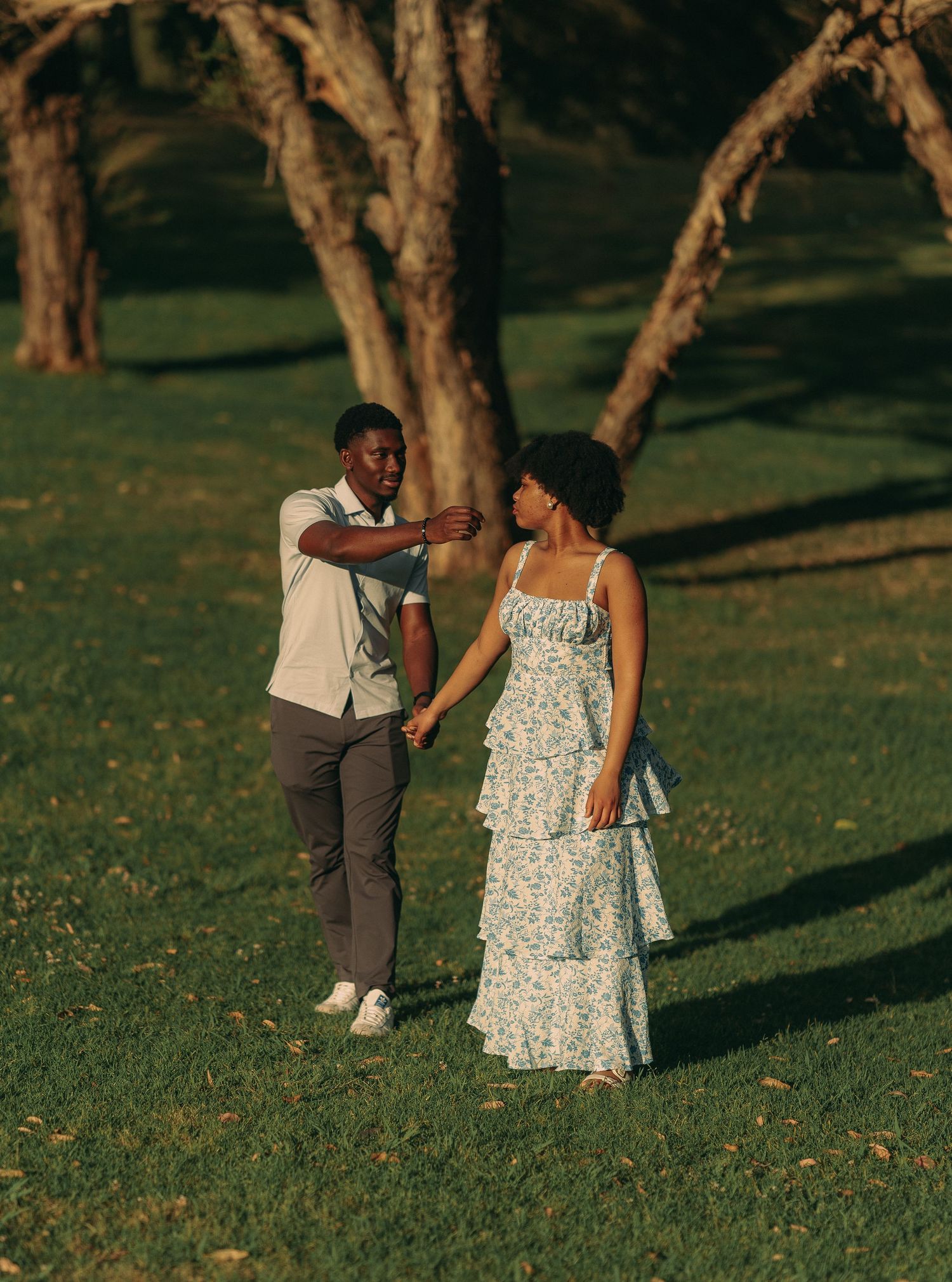 A romantic couple holds hands and dances together on a grassy park lawn surrounded by trees at sunset.