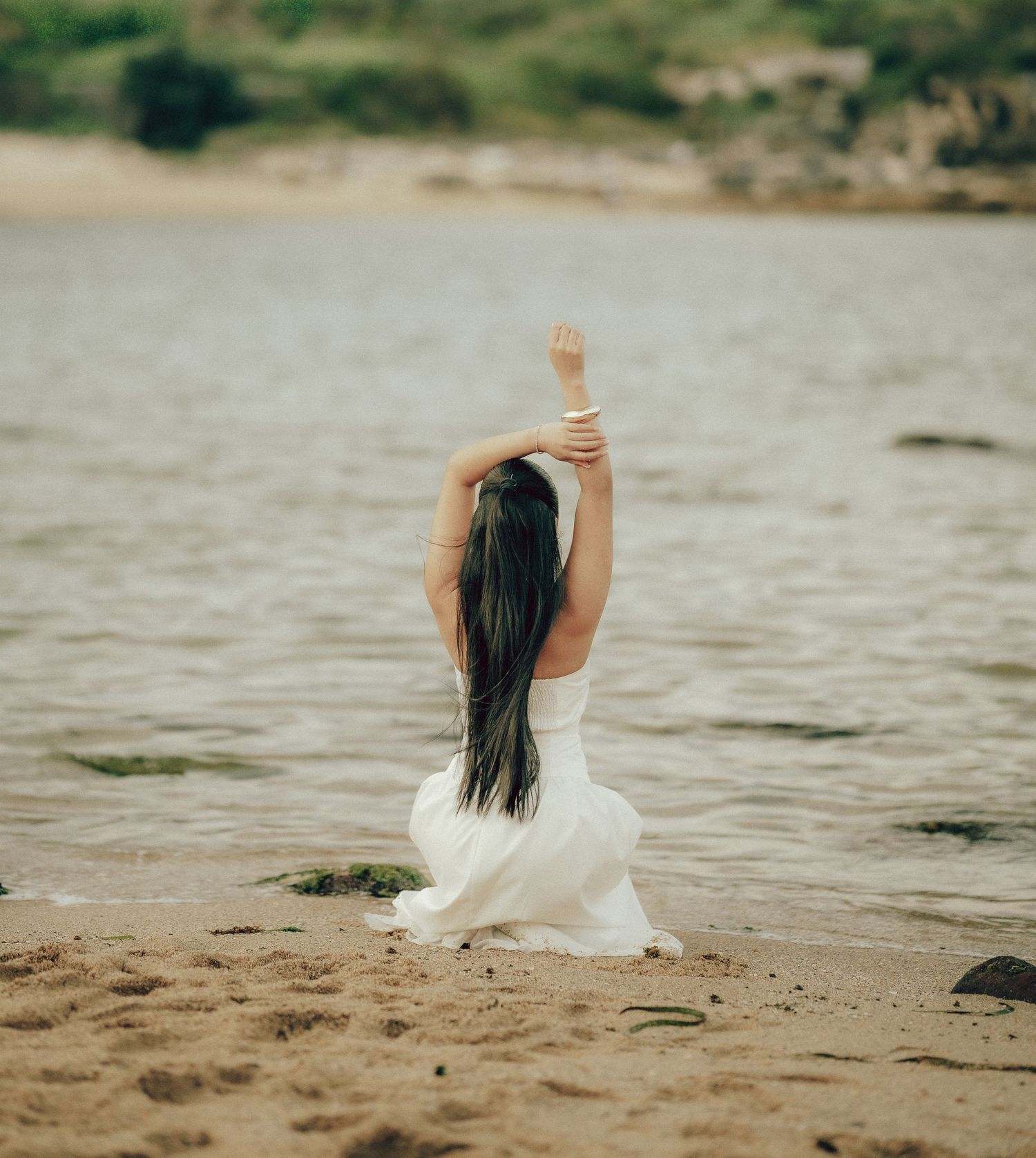 Woman in white dress sitting on sandy beach facing the water with arm raised gracefully overhead.