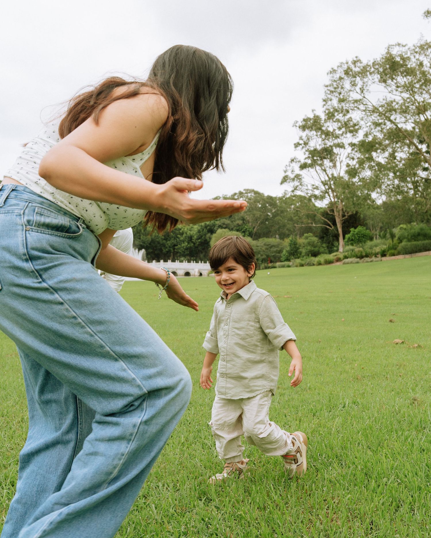 A playful mother reaches toward her giggling toddler son as they run together on a lush green park lawn.