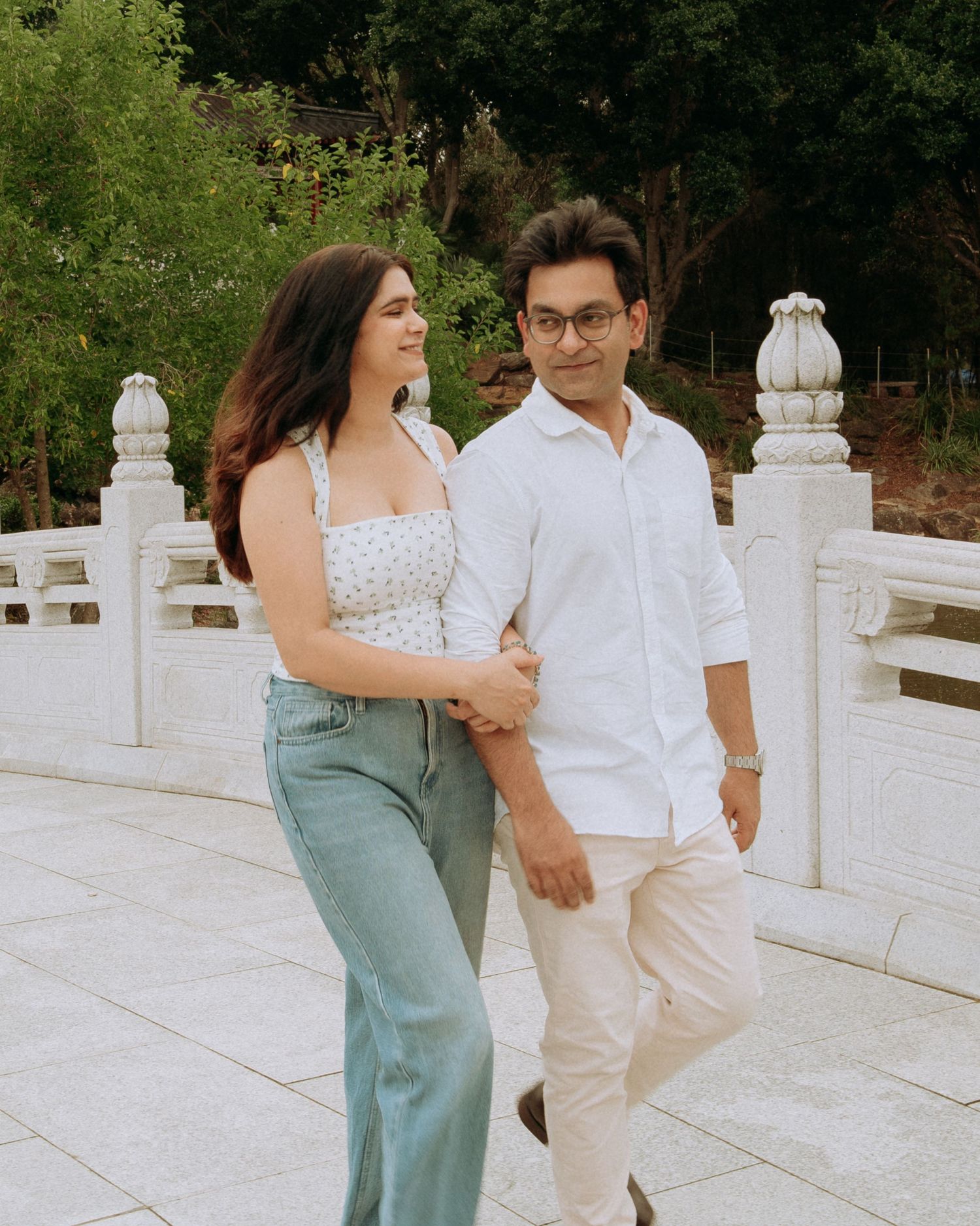 A stylish couple walks arm in arm on a white marble bridge surrounded by lush green gardens.
