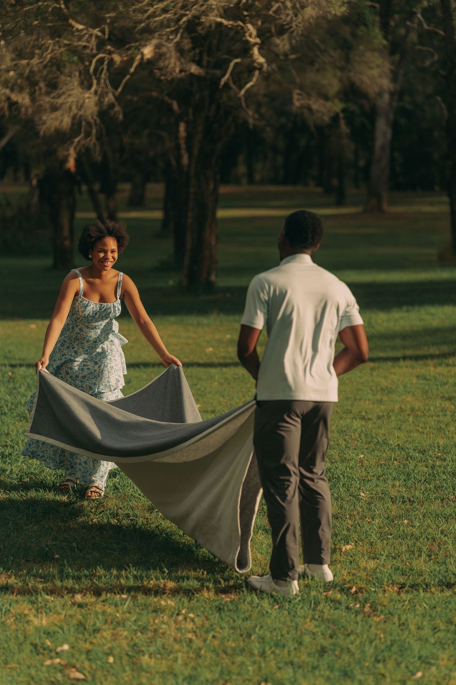 A smiling couple spreads a gray blanket together on a sunlit green lawn surrounded by trees.