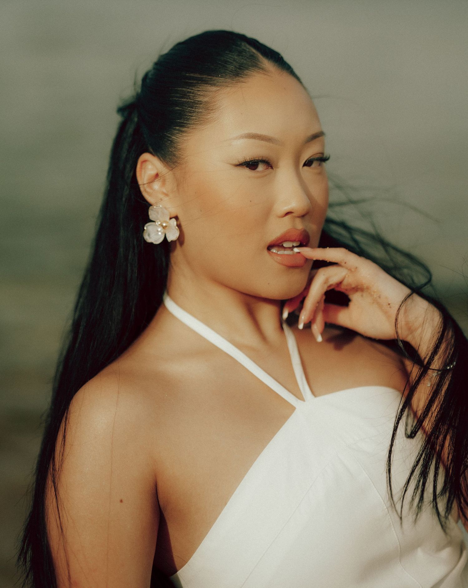 Close-up portrait of Asian woman in white halter top wearing floral earrings with windswept hair outdoors.