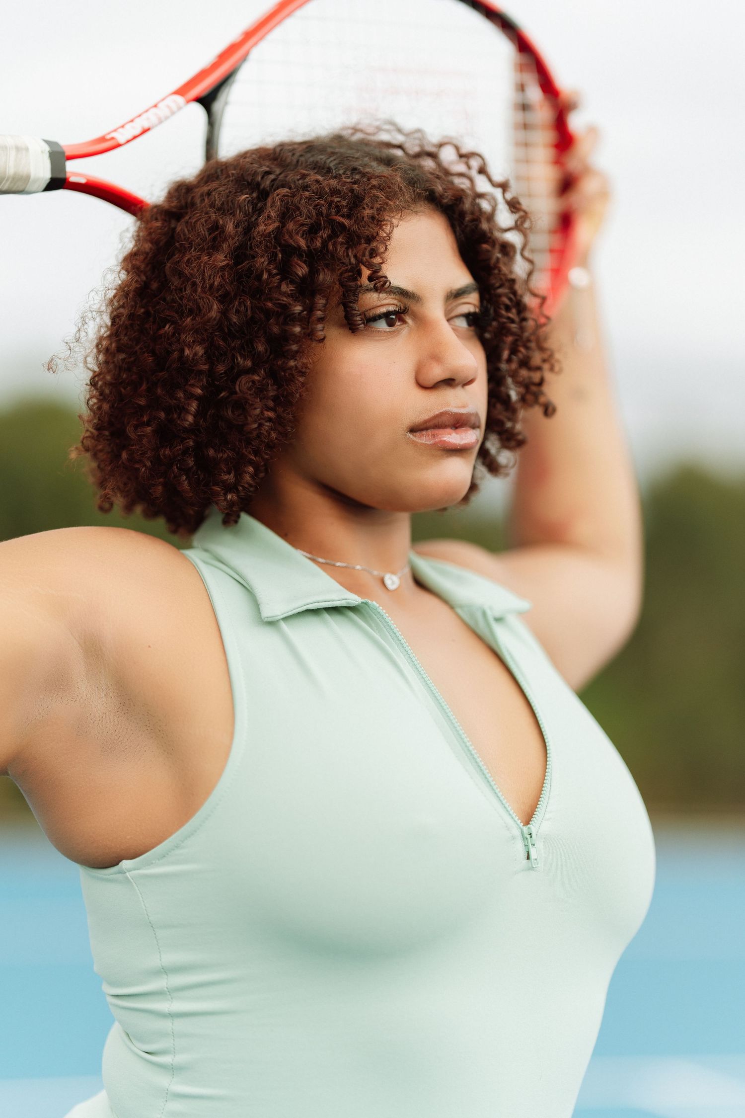 A curly-haired woman in a mint green tennis top holds a red racket above her head on an outdoor court.