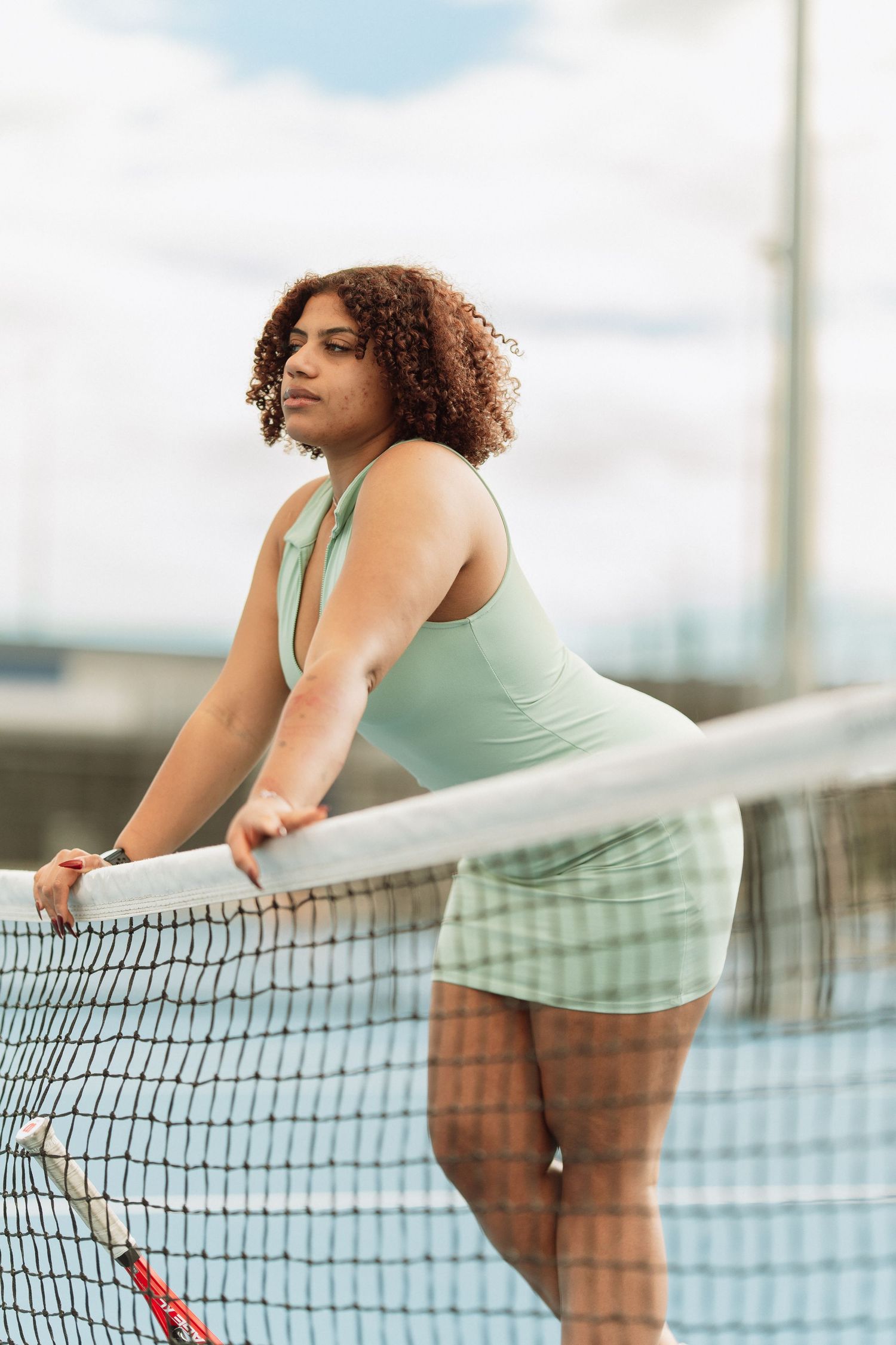 Woman in mint green athletic wear leans against tennis net on outdoor court holding a racket.