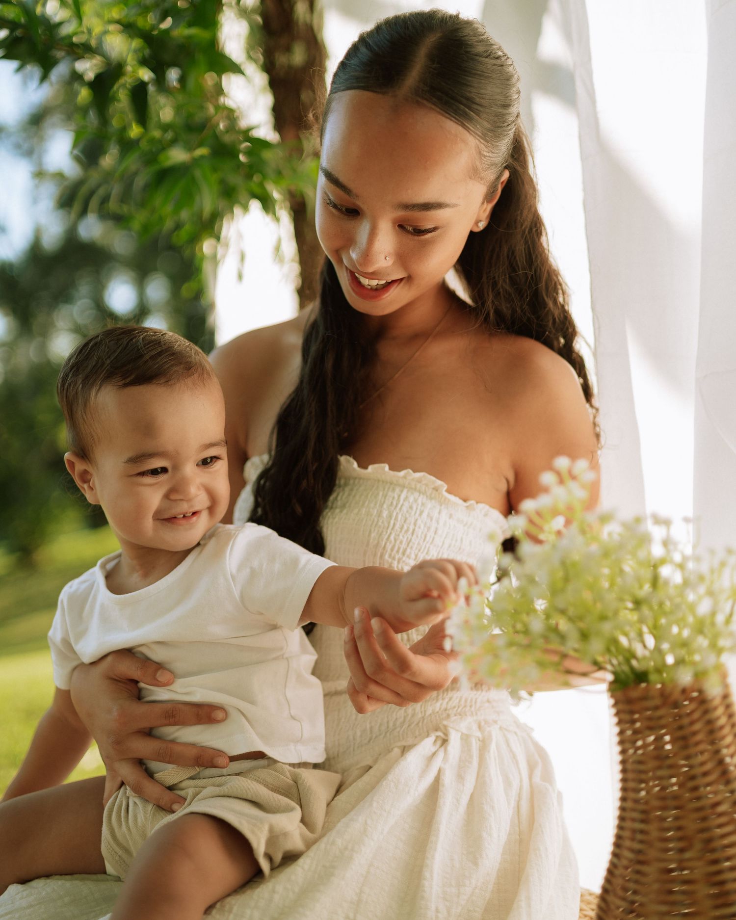 Young mother in white strapless dress sitting with smiling baby boy reaching toward flower basket in sunlit outdoor setting.