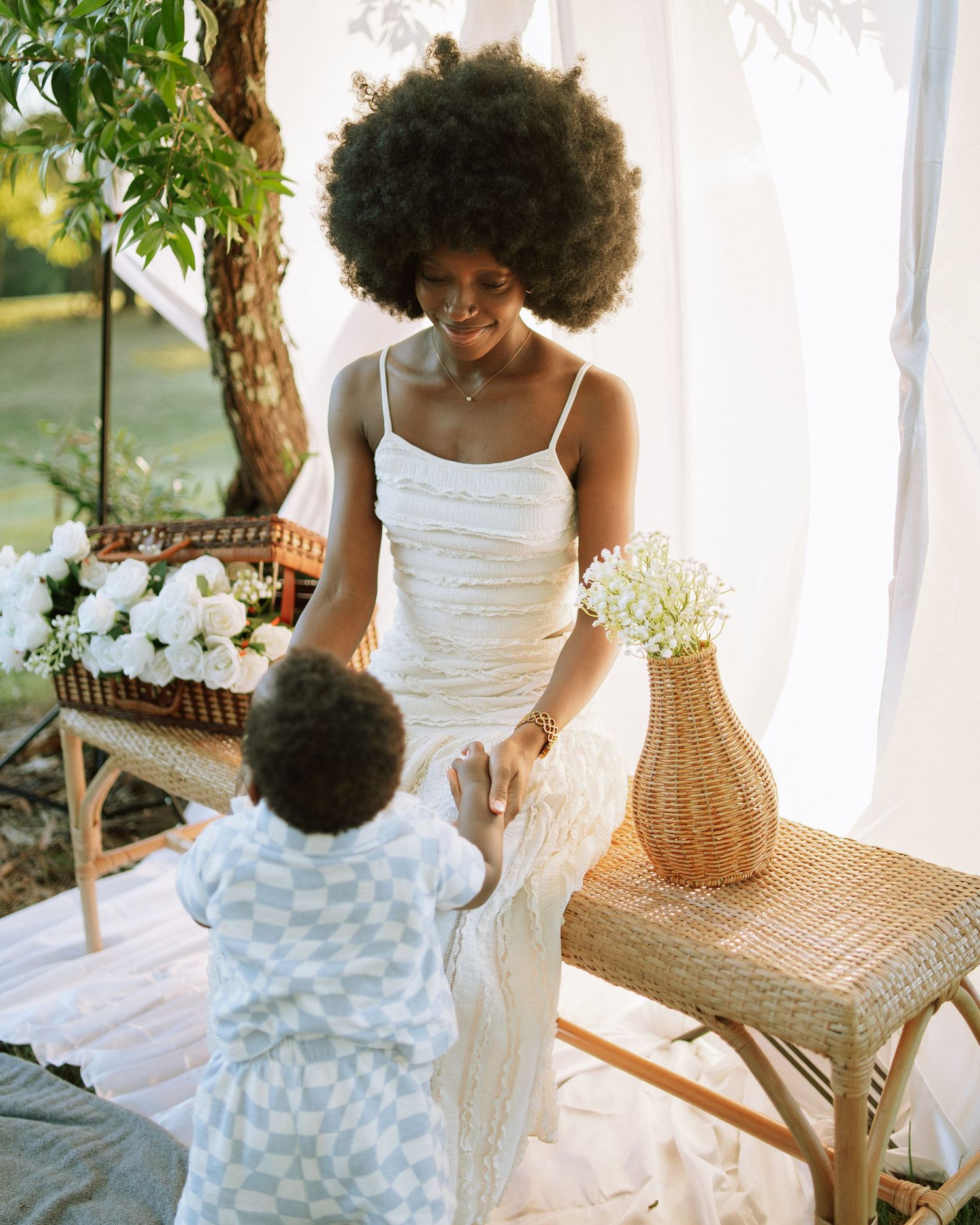 Elegant Black mother in white dress holding toddler son's hands at a beautifully styled outdoor setup.