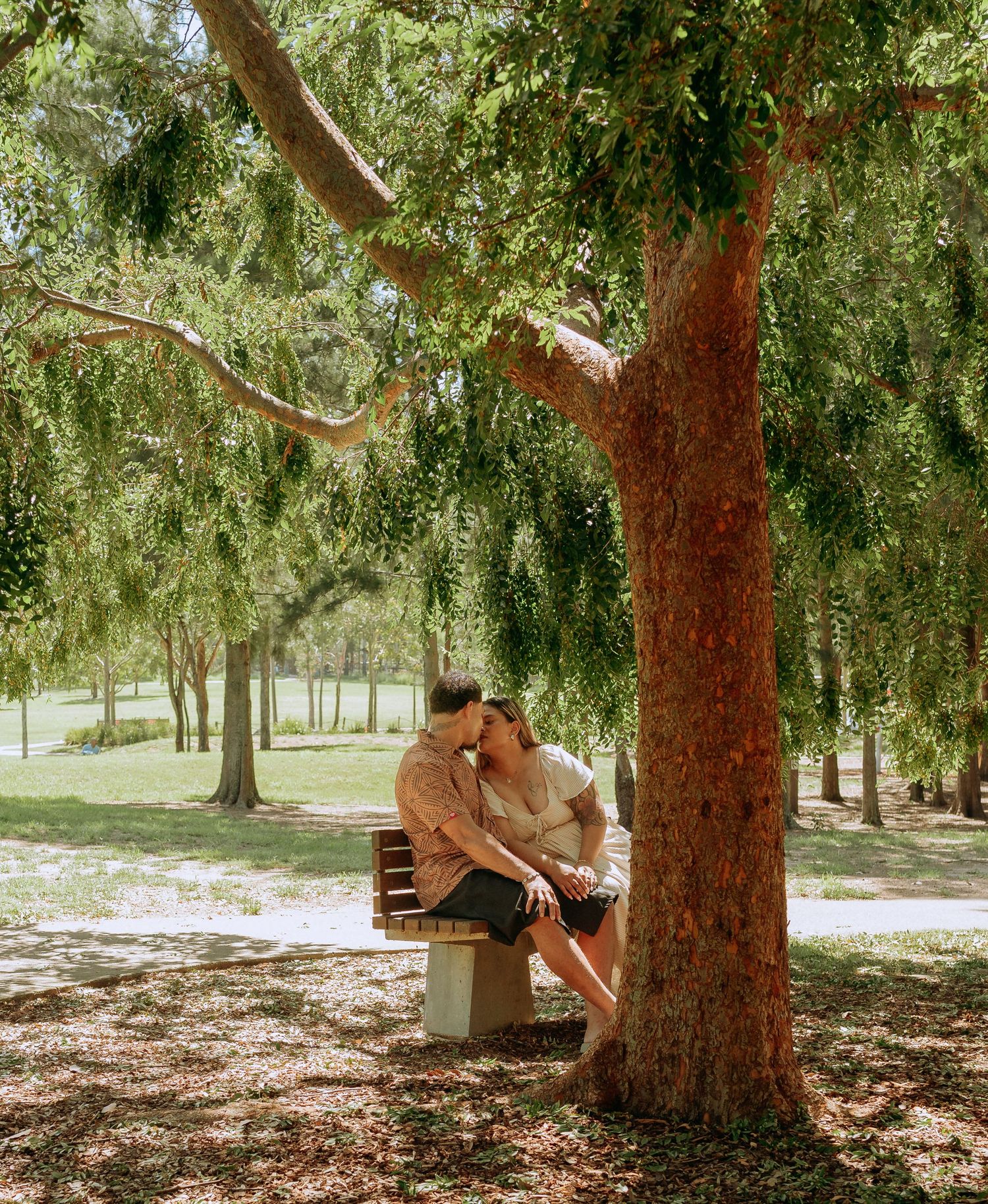 Romantic couple sharing a tender kiss while seated on a bench under a large shady tree in a park.