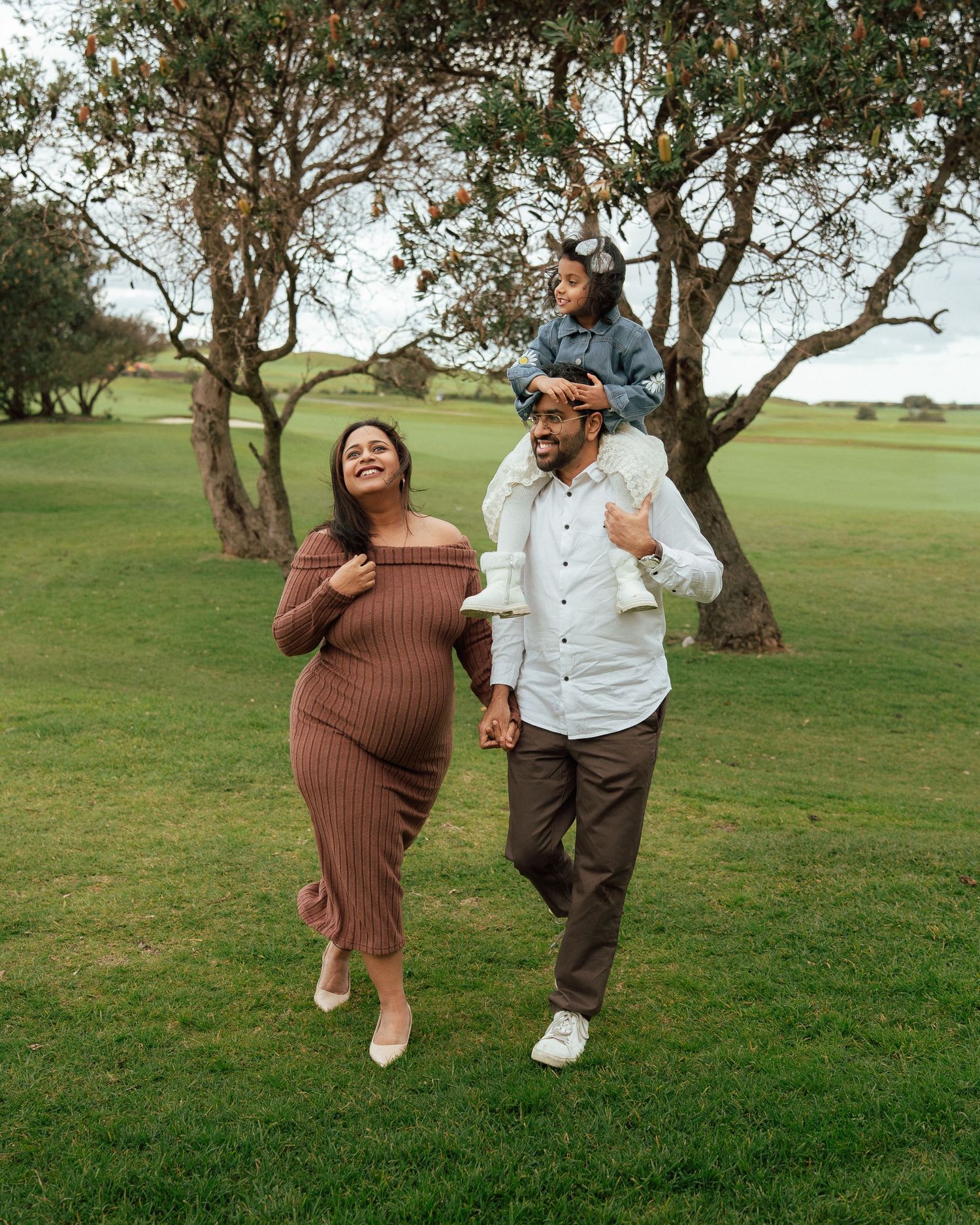 Pregnant woman in brown dress walks with man carrying child on shoulders on green golf course.