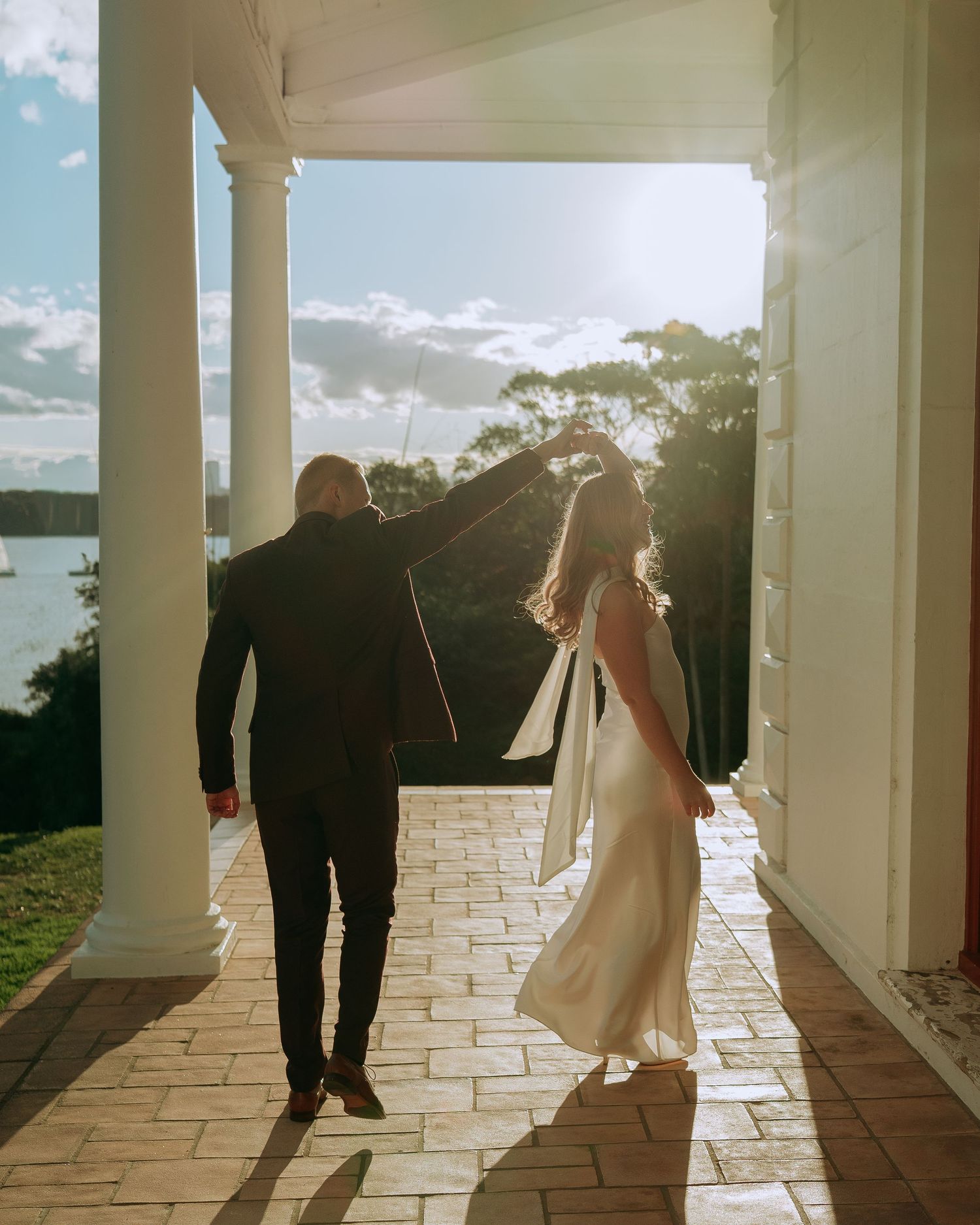 Couple in wedding attire walk hand-in-hand through sunlit colonial veranda overlooking waterfront.