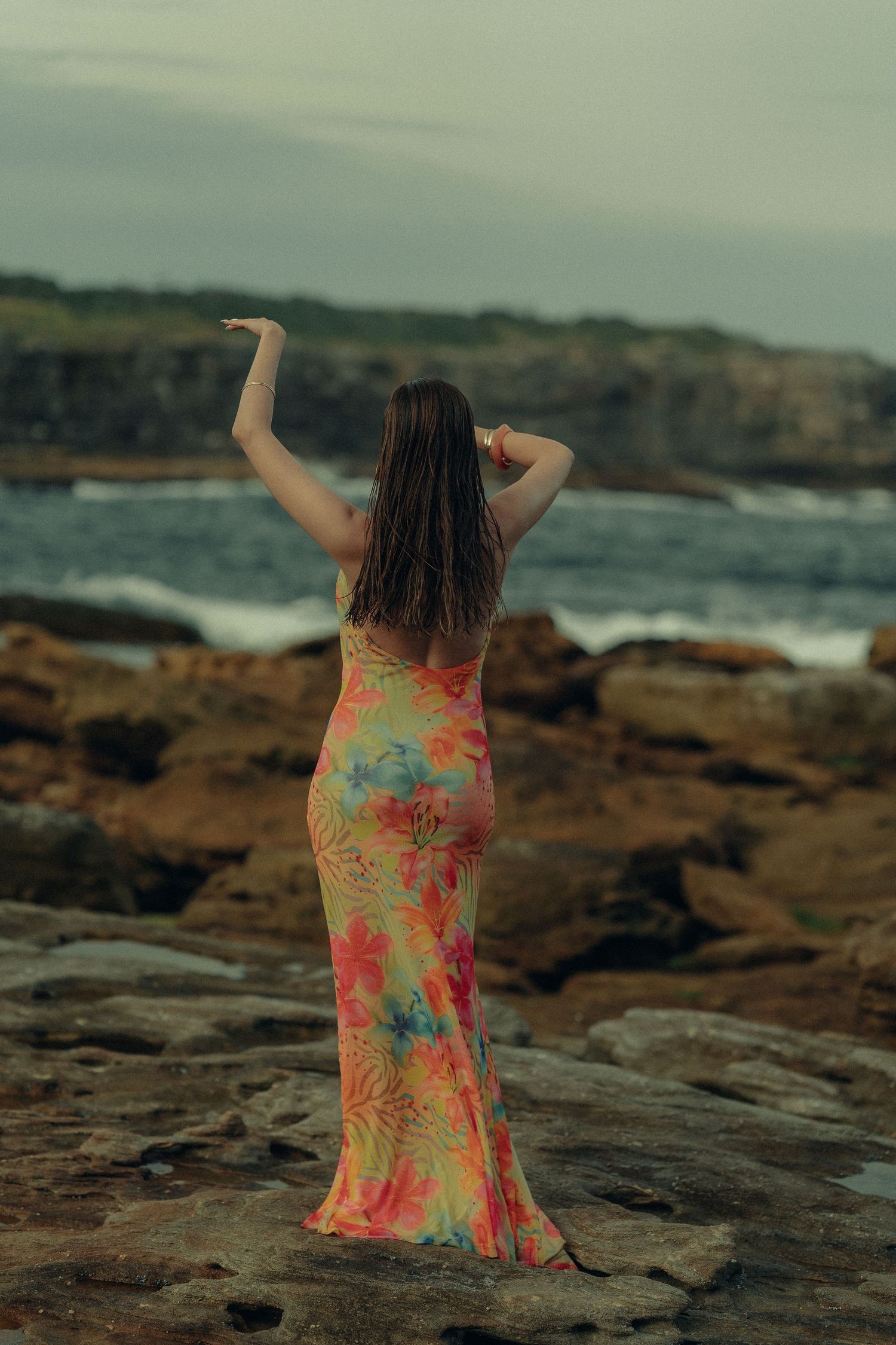 Woman in colorful tropical floral dress posing on rocky seaside cliffs with one arm raised upward.
