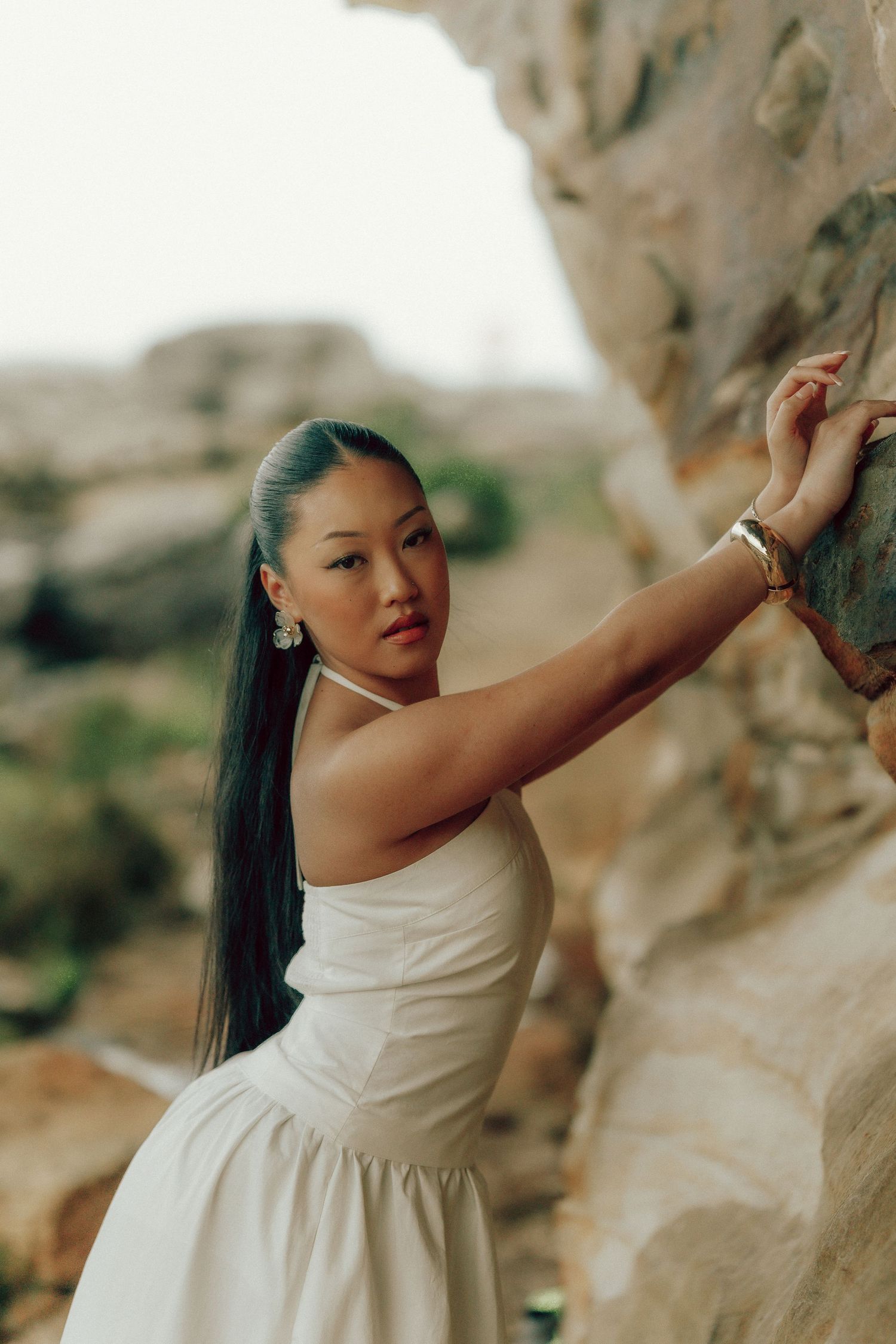 Asian woman in white dress touching rocky cliff face wearing floral earrings and bracelet outdoors.