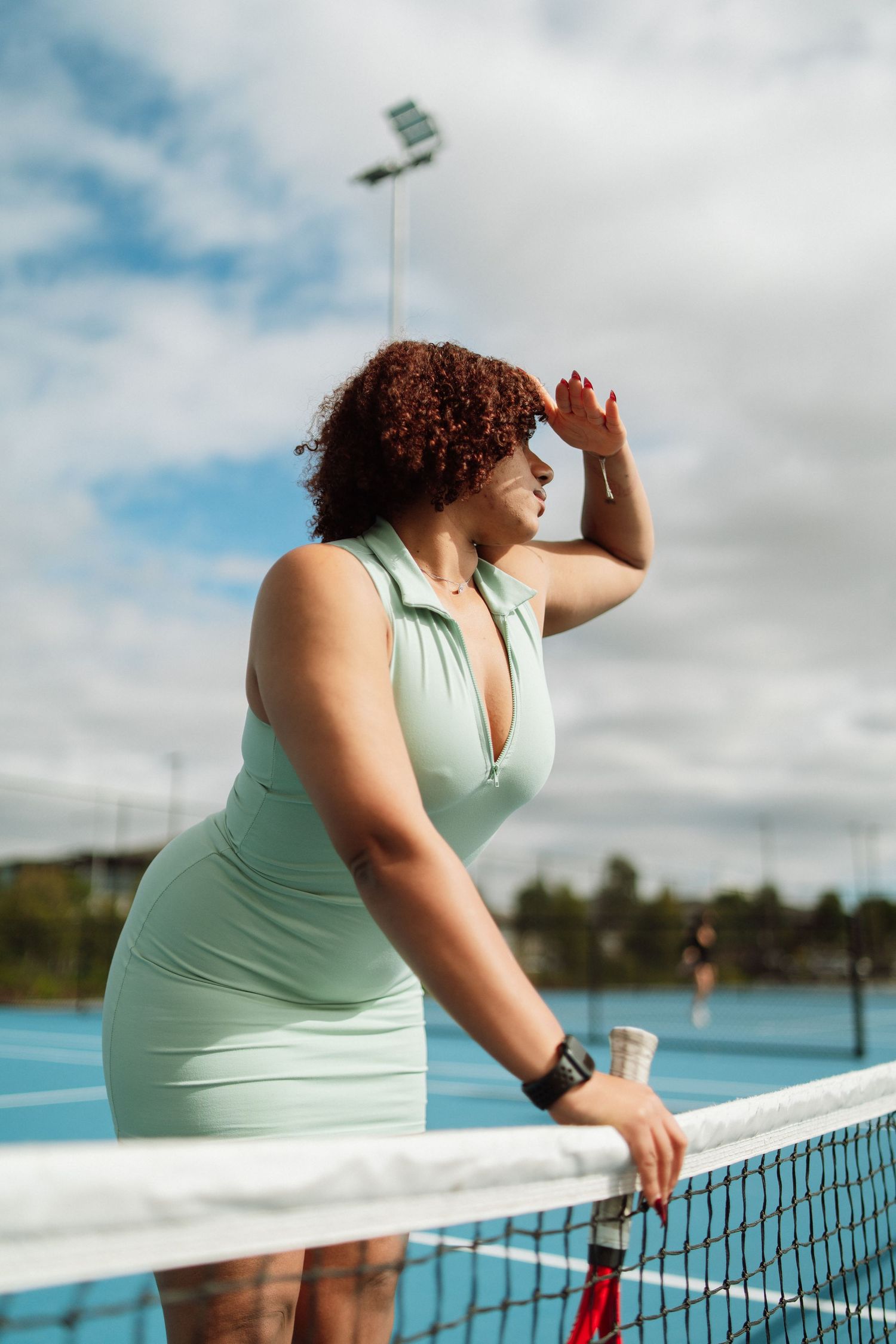 A woman in a mint green tennis dress shields her eyes while leaning on the net of a blue outdoor tennis court.