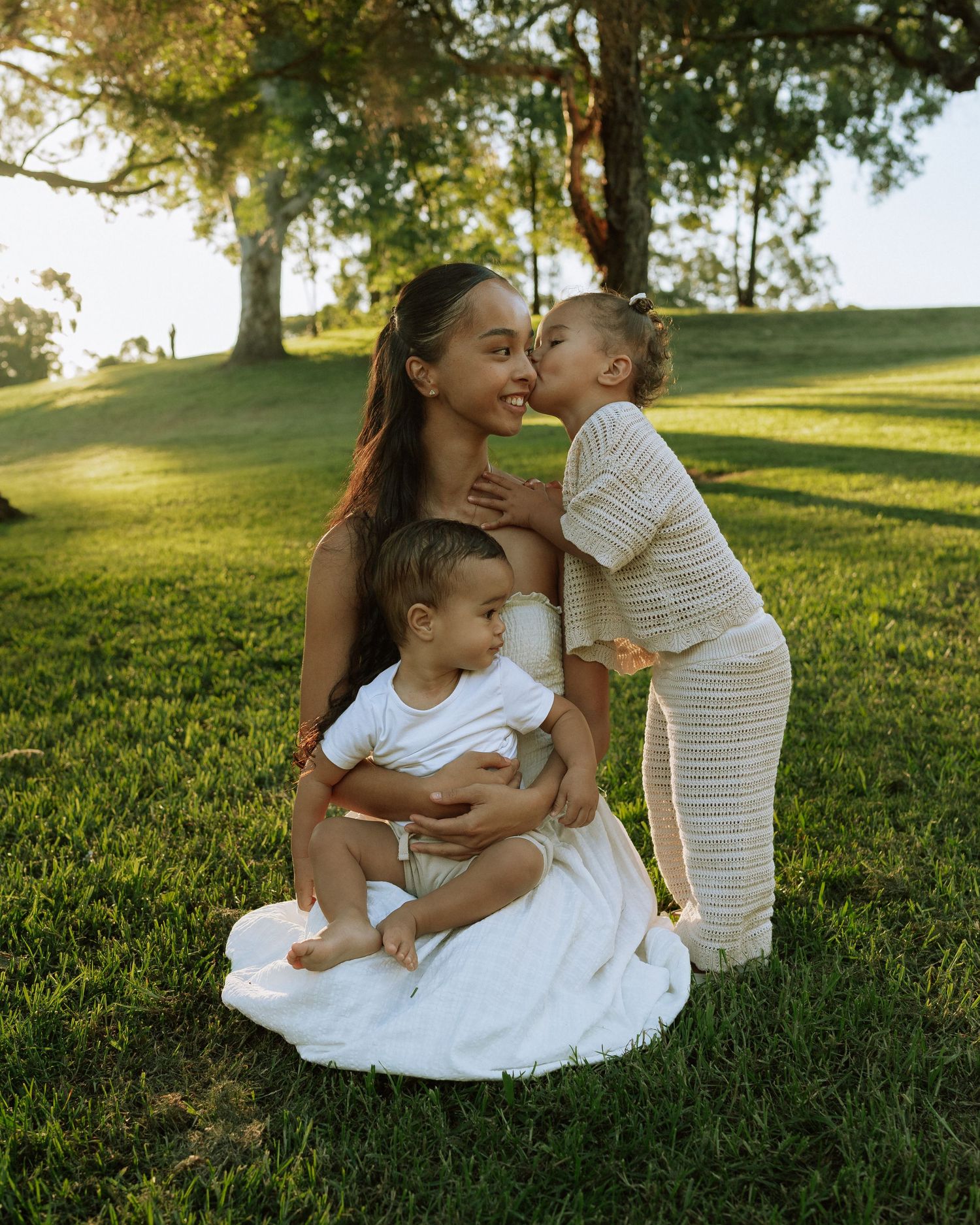 A mother kneels on green grass holding her baby while her toddler gives her a sweet kiss.