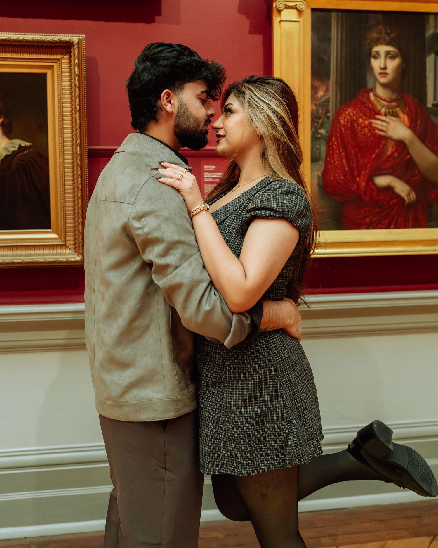 Romantic couple embracing and almost kissing in an art museum with classical paintings on red walls.