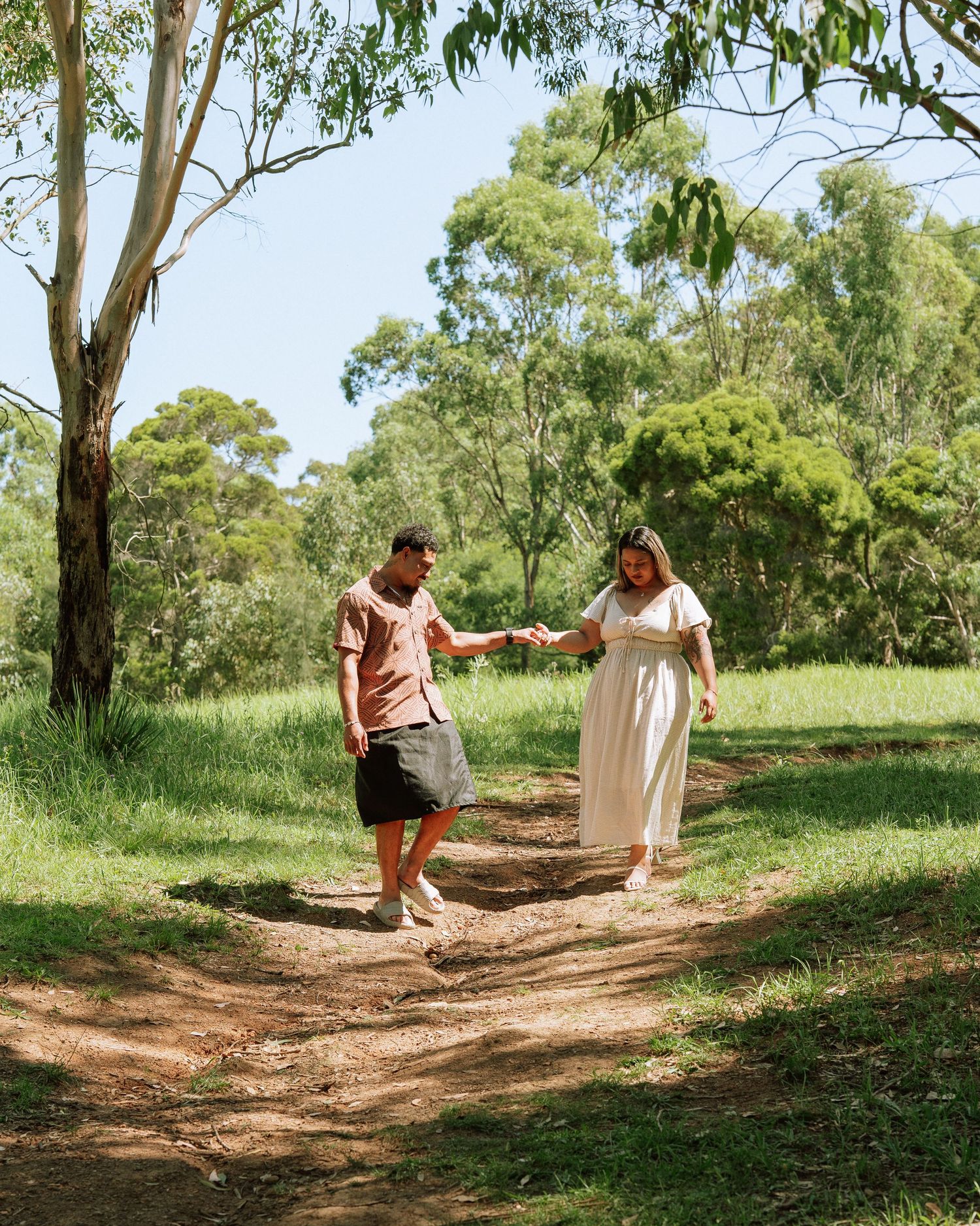 Couple holding hands walking on a dirt path surrounded by lush green trees and grass on a sunny day.