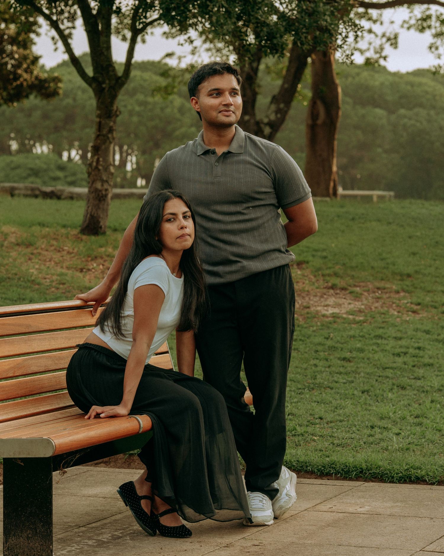 Young couple posing together in a park, woman sitting on bench while man stands beside her.