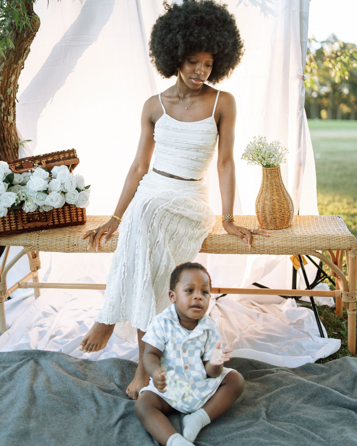 A mother with a natural afro in white outfit poses with her seated baby during an elegant outdoor photoshoot.
