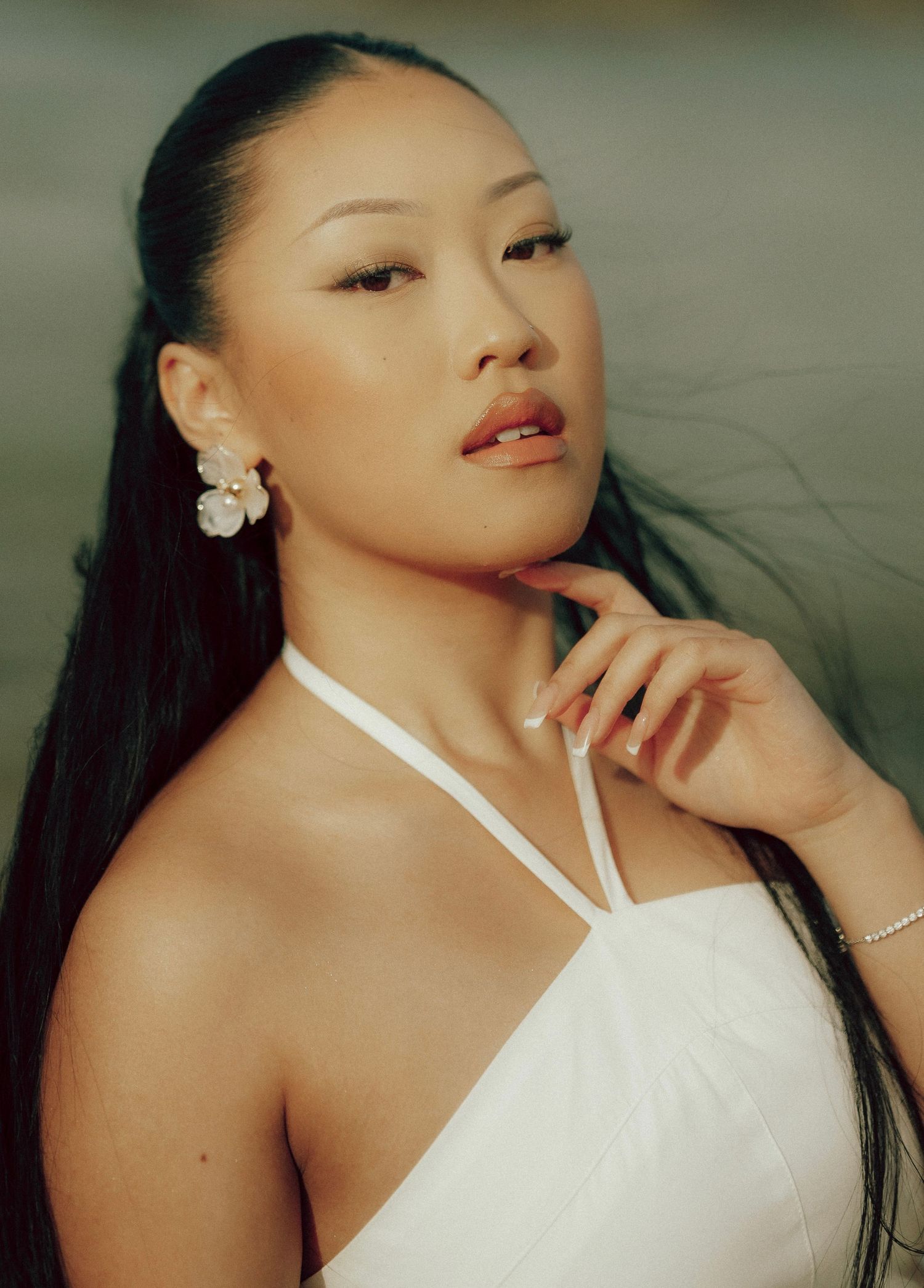 Close-up portrait of Asian woman with sleek hair wearing white halter top and floral earrings outdoors.