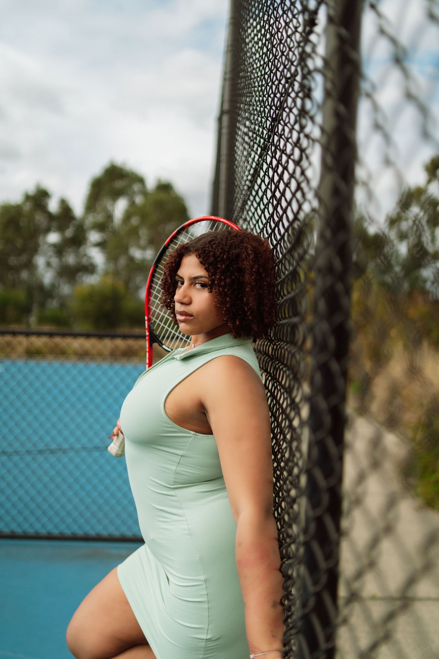 Athletic woman in mint green dress leaning against tennis court fence holding racket.