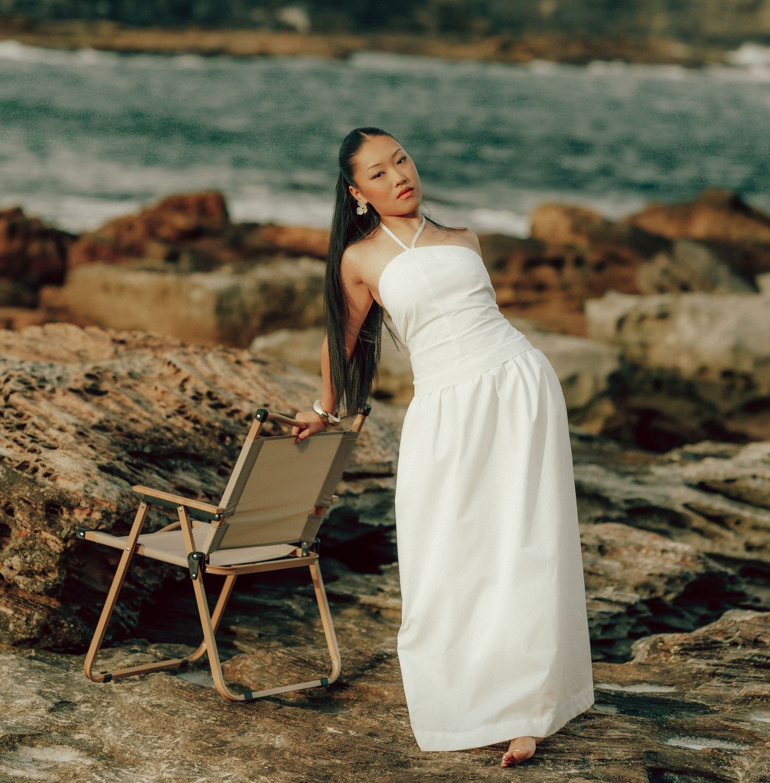 Woman in white halter dress posing on rocky coastline beside a folding chair with ocean waves behind.