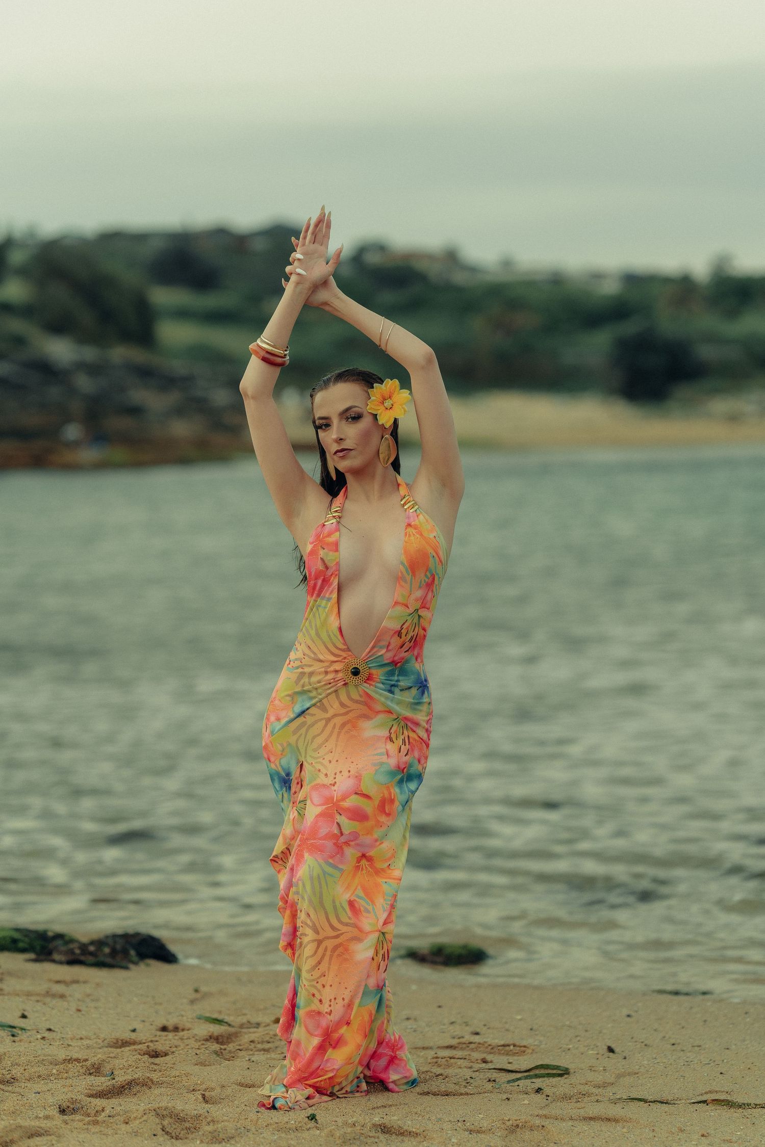 Woman in bright floral halter dress stands on sandy beach with arms raised and yellow flower in hair.