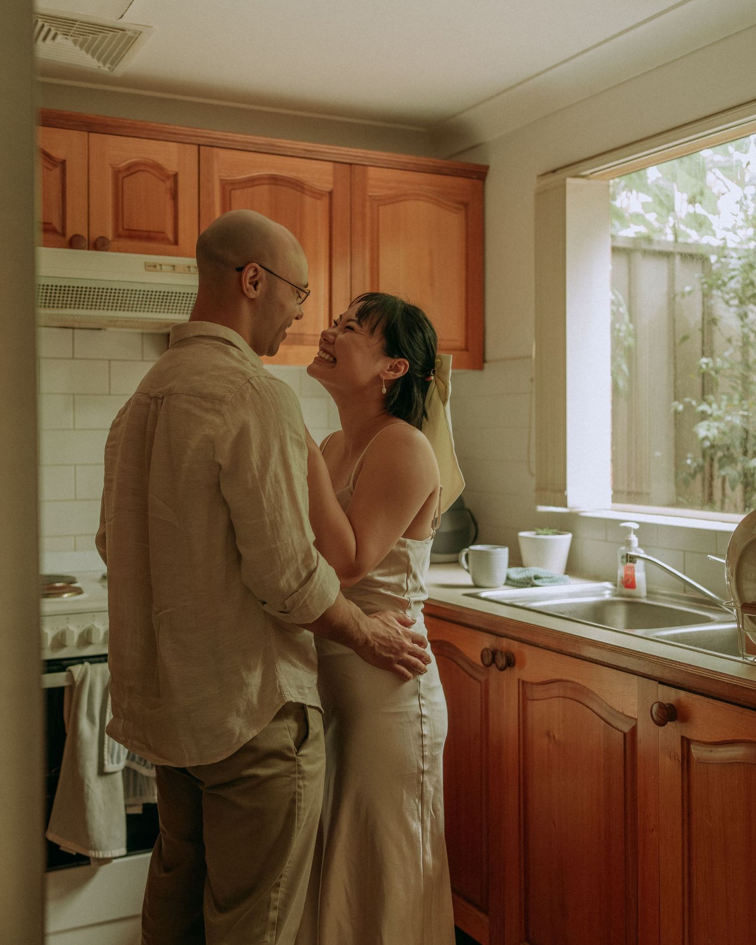 Couple laughing and embracing intimately in a warm wooden kitchen near a window with natural light.