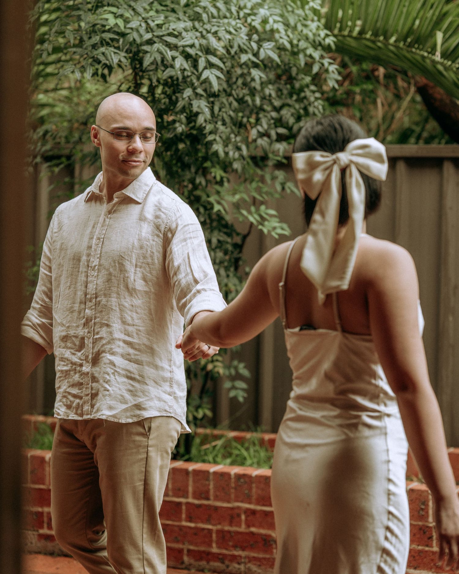 Groom in linen shirt holds bride's hand as she leads him through a lush brick garden.