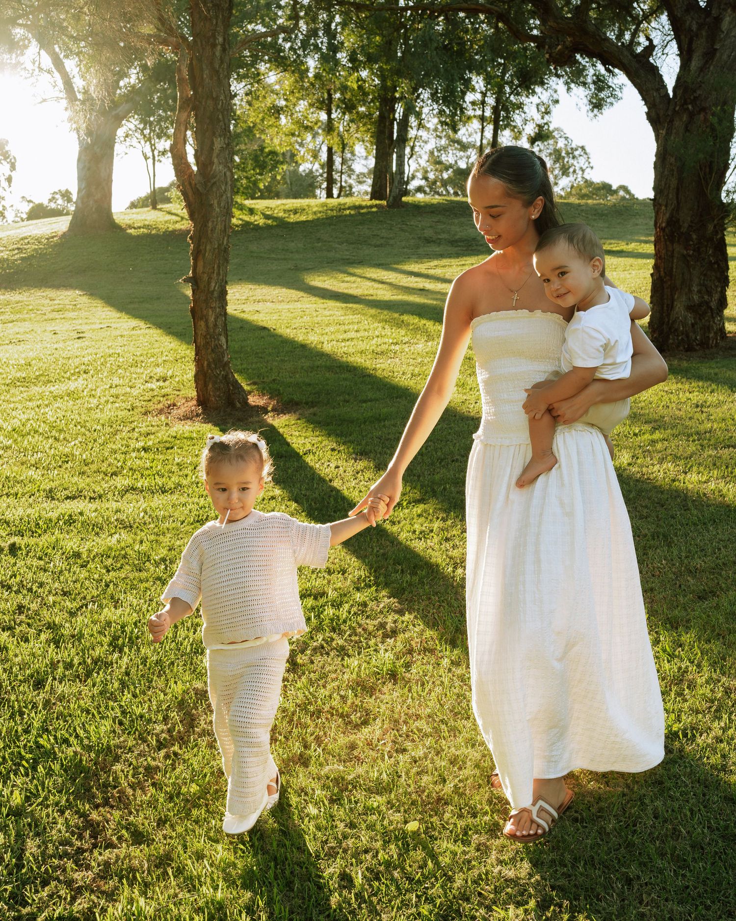 Woman in white dress holding baby and toddler's hand on sunny green lawn with trees.